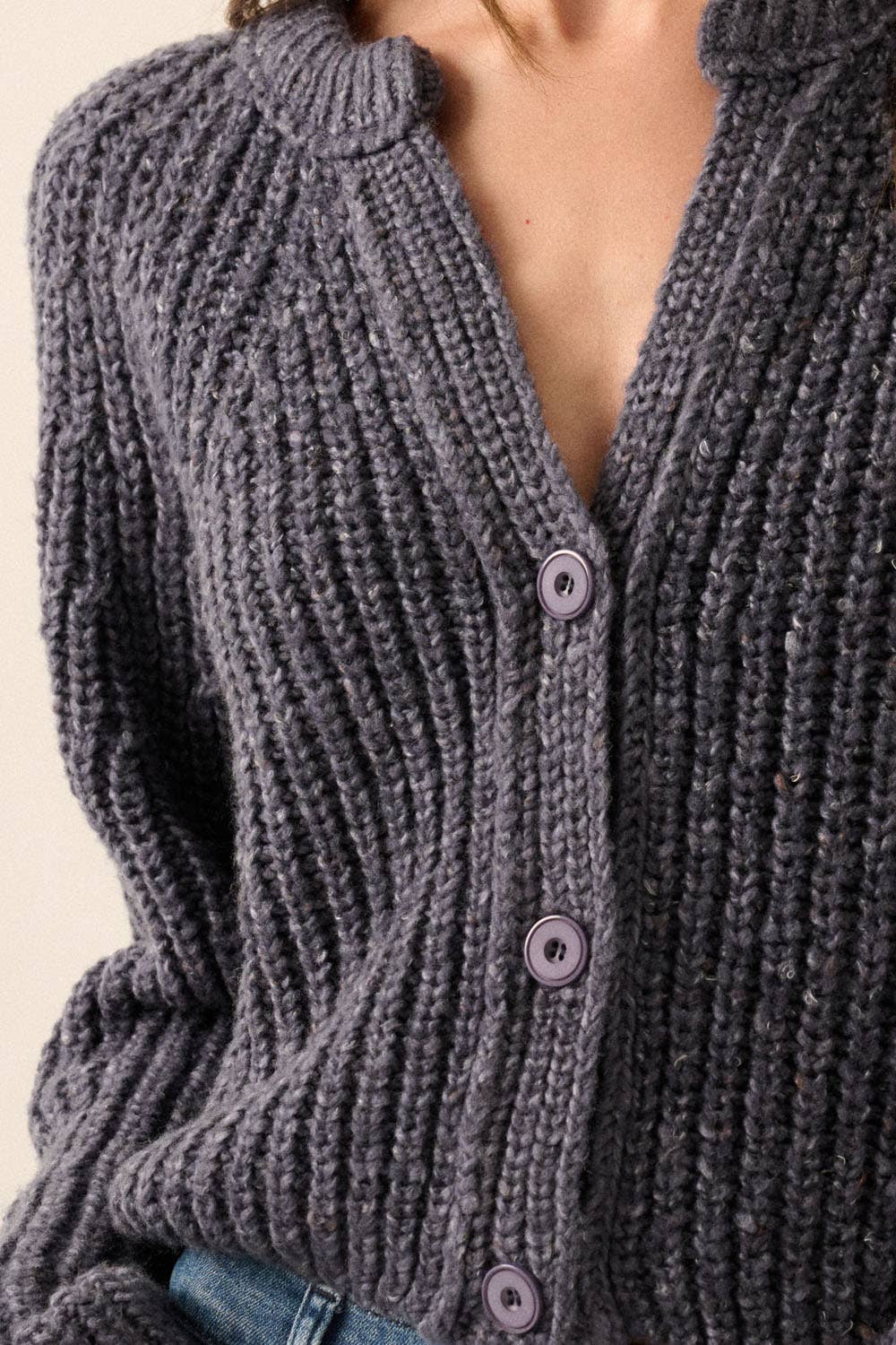 A person wears the Promesa USA Confetti Button-Front Cardigan Sweater in dark gray with three light purple buttons, partially unbuttoned, paired with blue jeans. The cardigan is the focus, and the face is not visible in the image.