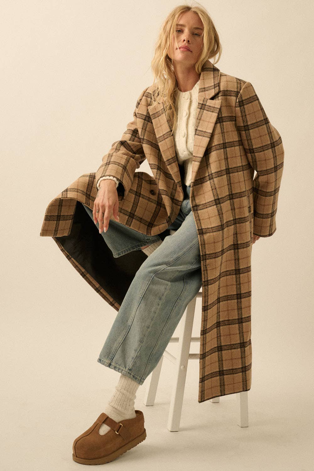 A woman with long blonde hair sits on a white stool against a plain background, wearing the Plaid Felt Double-Breasted Calf-Length Overcoat by Promesa USA, a white textured sweater, light blue wide-leg jeans, white socks, and brown slip-on shoes.