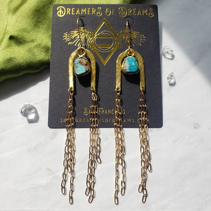 The Gold Rainbow Arch Chain Duster Earrings by Dreamers of Dreams feature turquoise stones and long gold-filled chain accents, displayed on a branded card with decorative stones scattered across a marble surface.