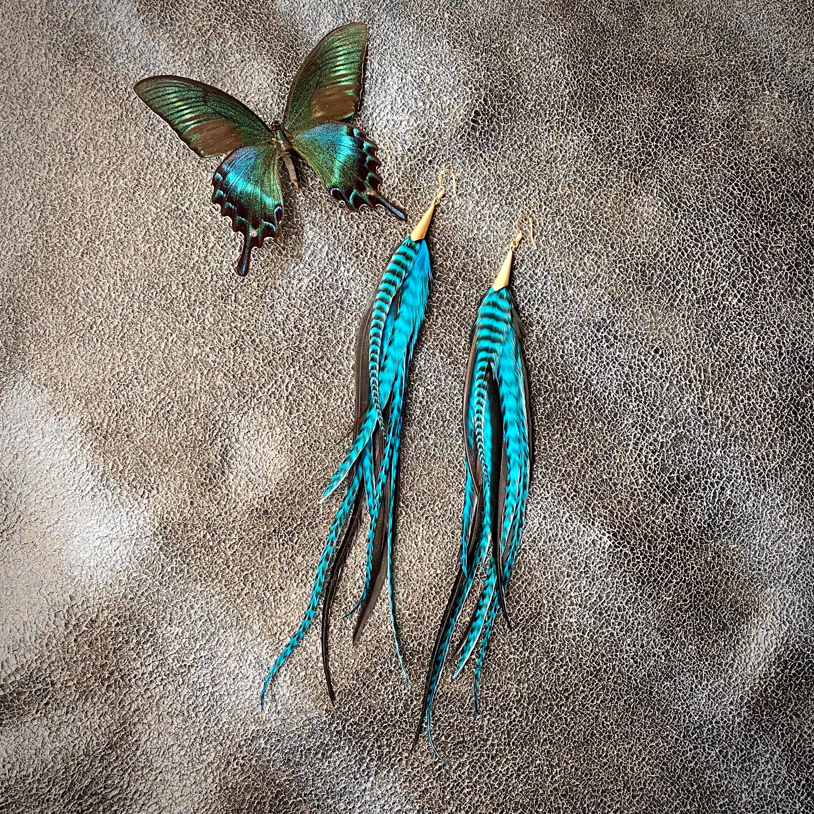 ASTALI’s Long Feather & Cone Earrings in Turquoise Grizzly, Black, and Gold are displayed alongside a green butterfly on a textured light brown surface. Earrings feature sustainably sourced feathers and gold hooks.