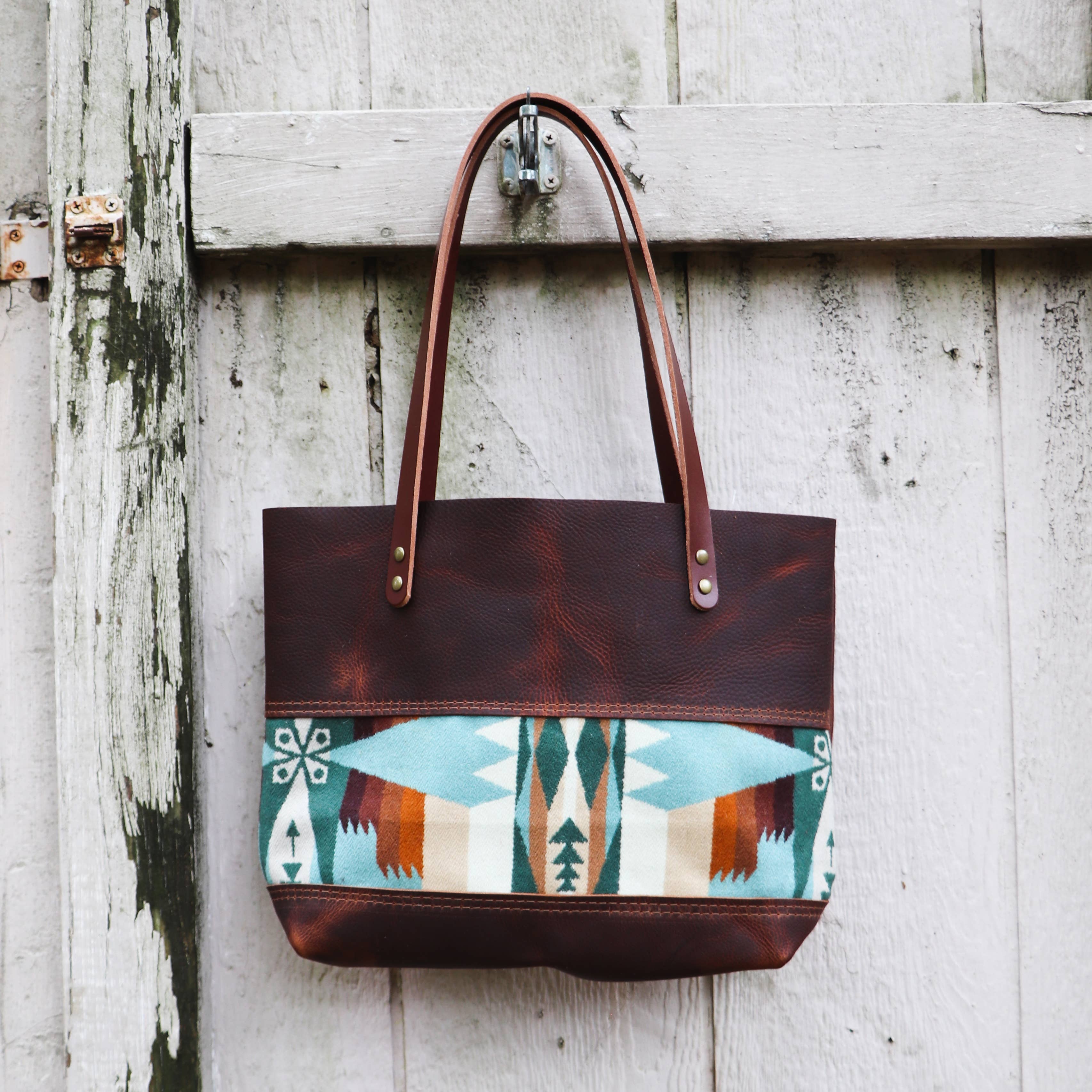 The Hawks & Doves Wool Tote Short features brown full grain leather, long handles, and a colorful geometric panel in teal, white, and orange, shown hanging on a weathered wooden wall.