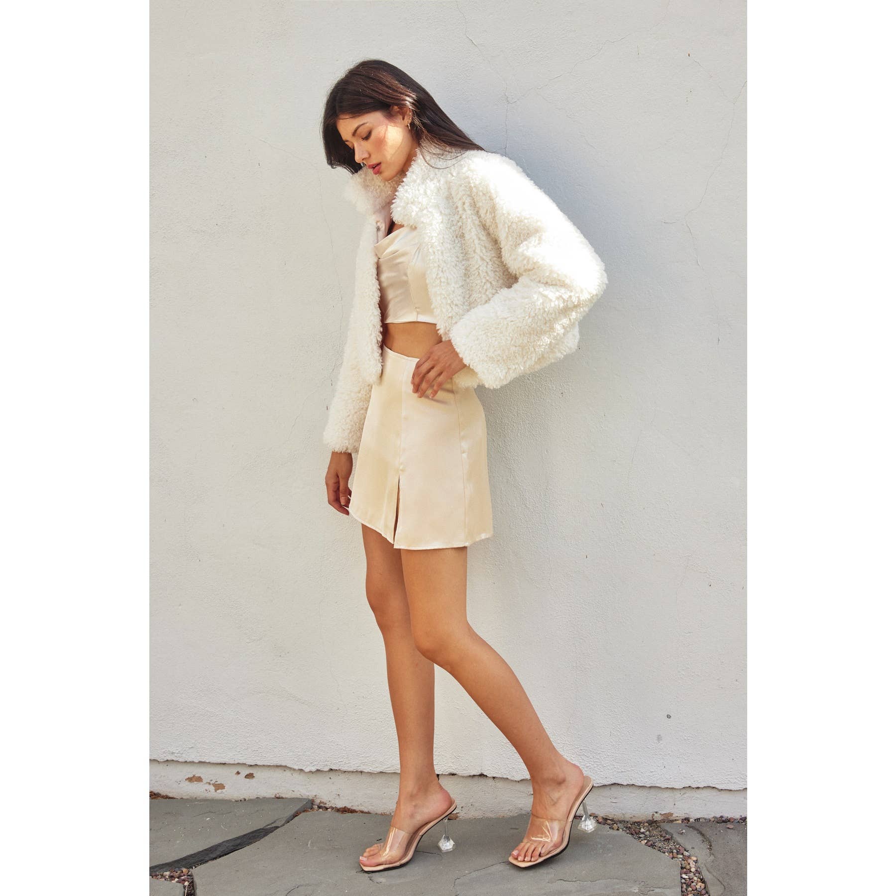 A woman stands against a white wall, wearing the Dress Forum Soft Teddy Open Front Cropped Jacket over a satin crop top and matching mini skirt, paired with clear high-heeled sandals. She looks down with one hand by her side.