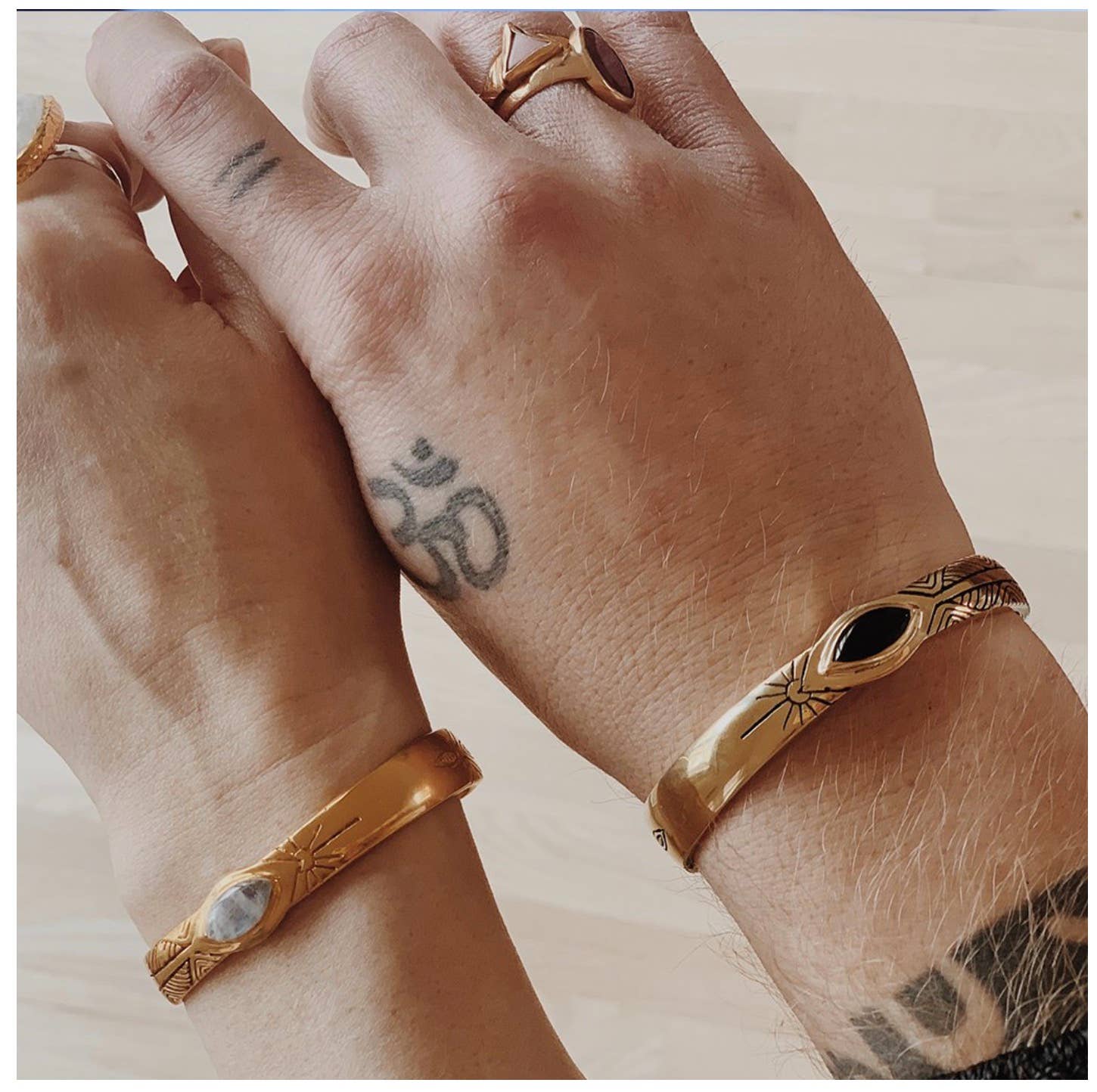Two hands wearing gold Three Rivers Cuff bracelets featuring artisan engraved designs and stones