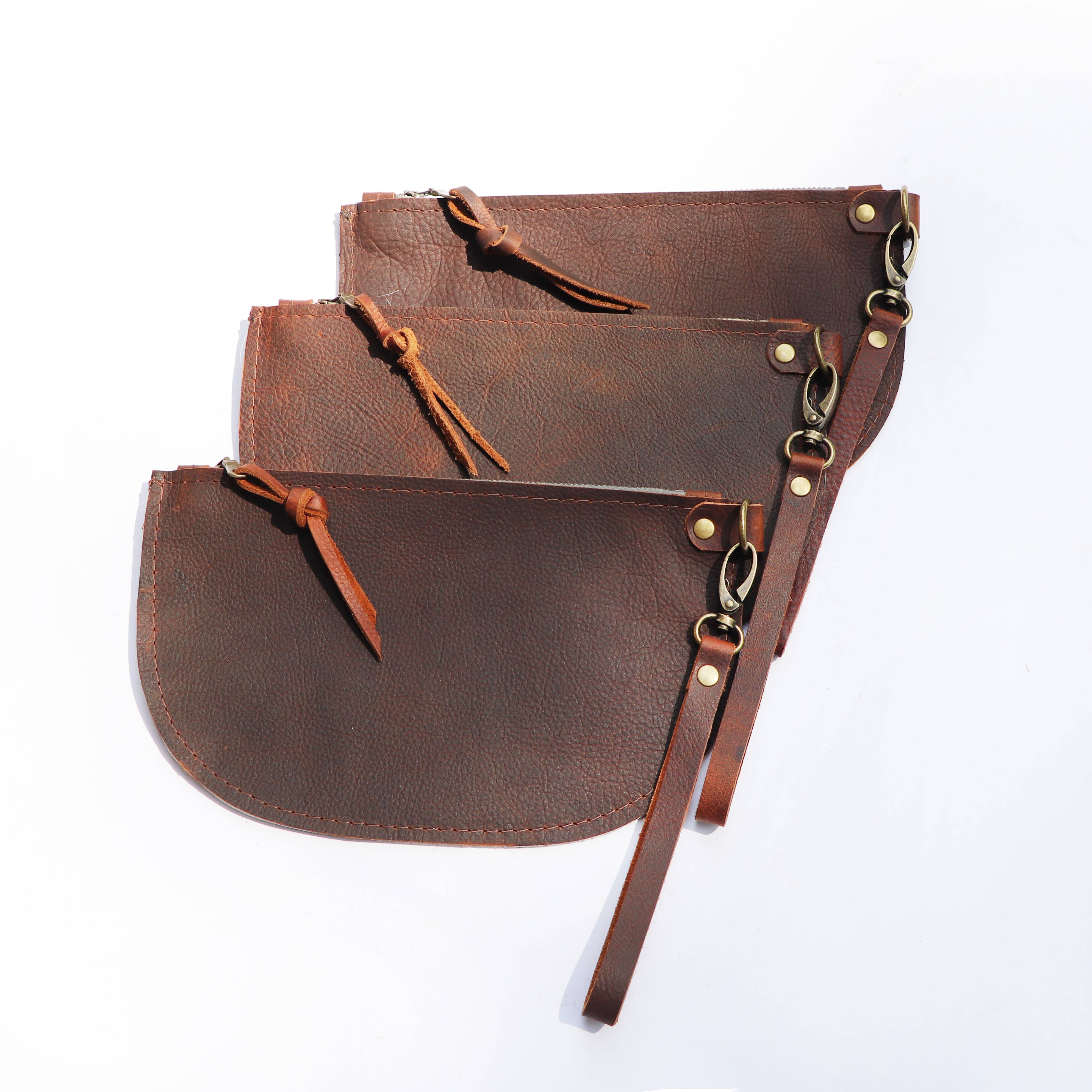 The Hawks & Doves Large Half Moon Cowhide and Leather Zip Pouch with Wristlet is shown stacked with other brown cowhide pouches, each featuring detachable wristlets attached by metal rings, displayed on a white background.