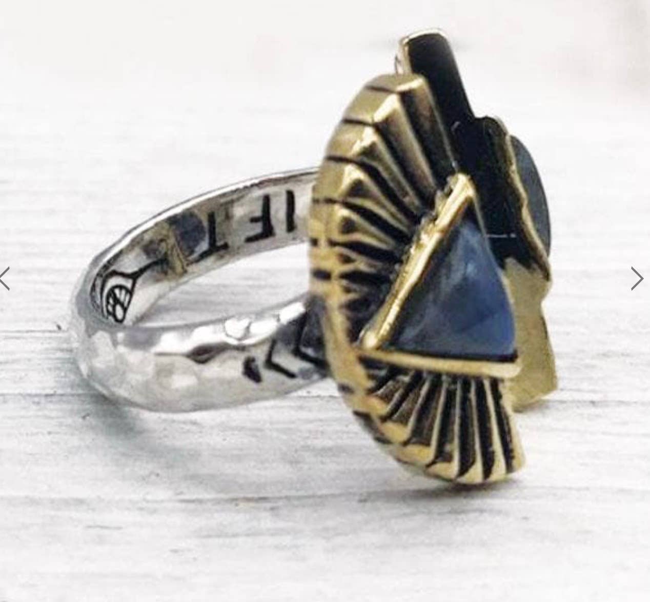 The Shape Shift Ring by bahgsu jewels is a sterling silver talisman with engraved details, showcasing a large triangular blue stone set in a gold fan-shaped design on top, displayed on a light surface.