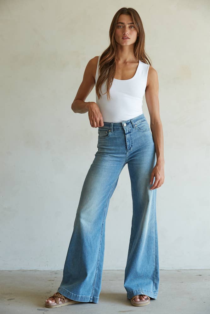 A woman with long brown hair poses confidently against a plain light wall, wearing By Together’s white Double Layered Seamless Top, high-waisted light blue flared jeans, and sandals.