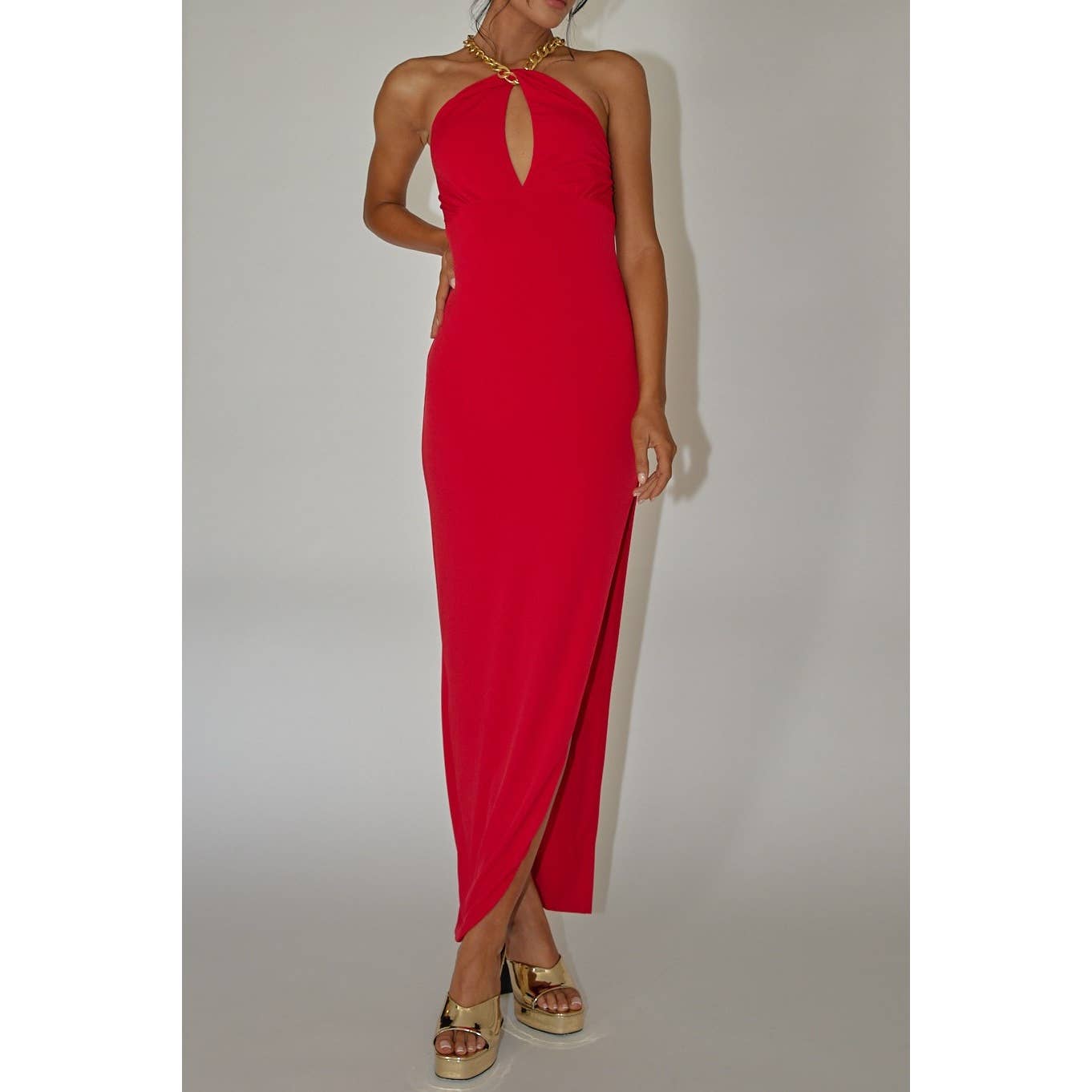 A woman wears the One and Only Collective Halterneck Gold Chain Red Maxi Dress, featuring a keyhole neckline and high slit, paired with gold platform sandals against a plain light background—an elegant evening look.