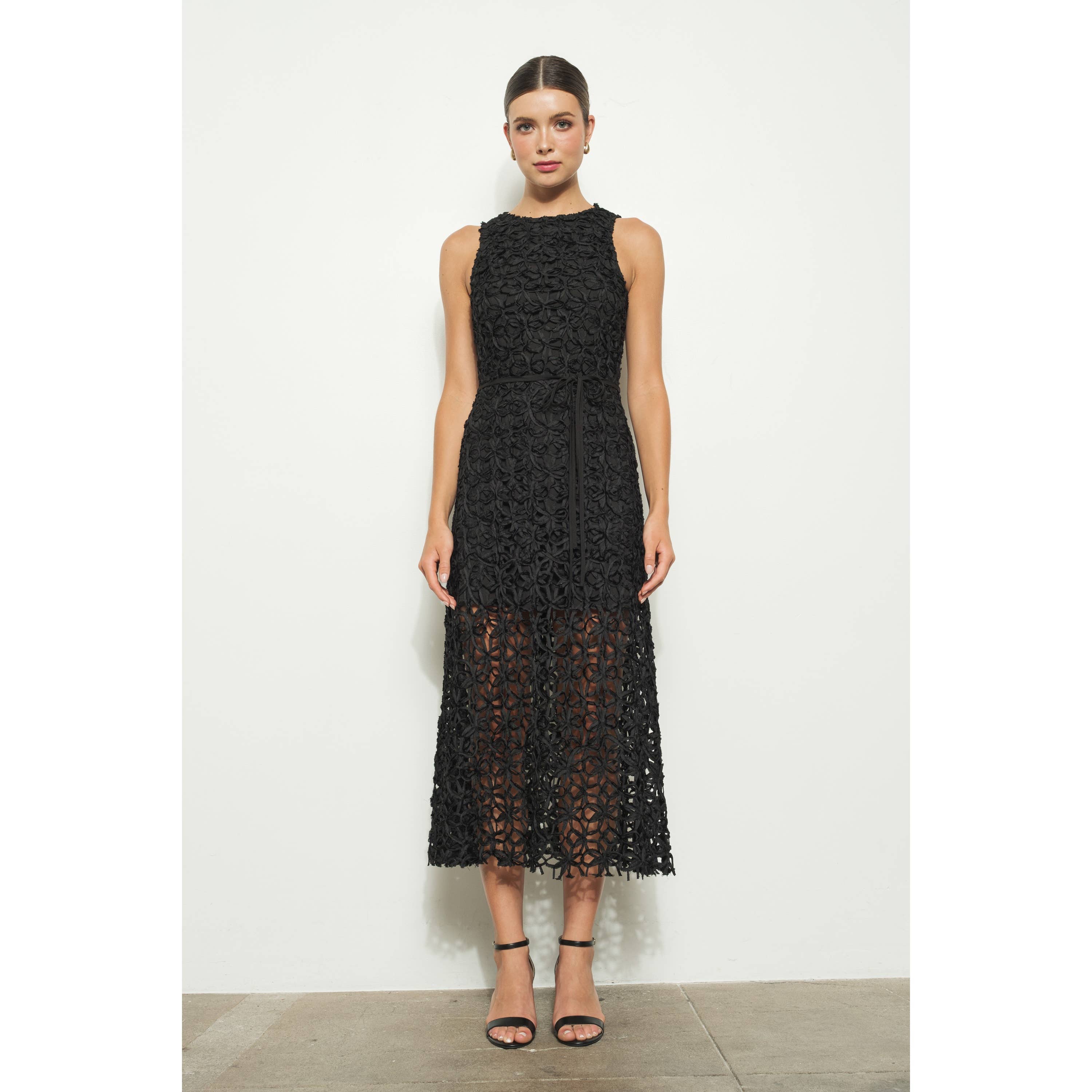 A woman stands against a white wall wearing the INA Sleeveless Crochet Lace Overlay Midi Dress with a fitted bodice and sheer skirt, paired with black high-heeled sandals.