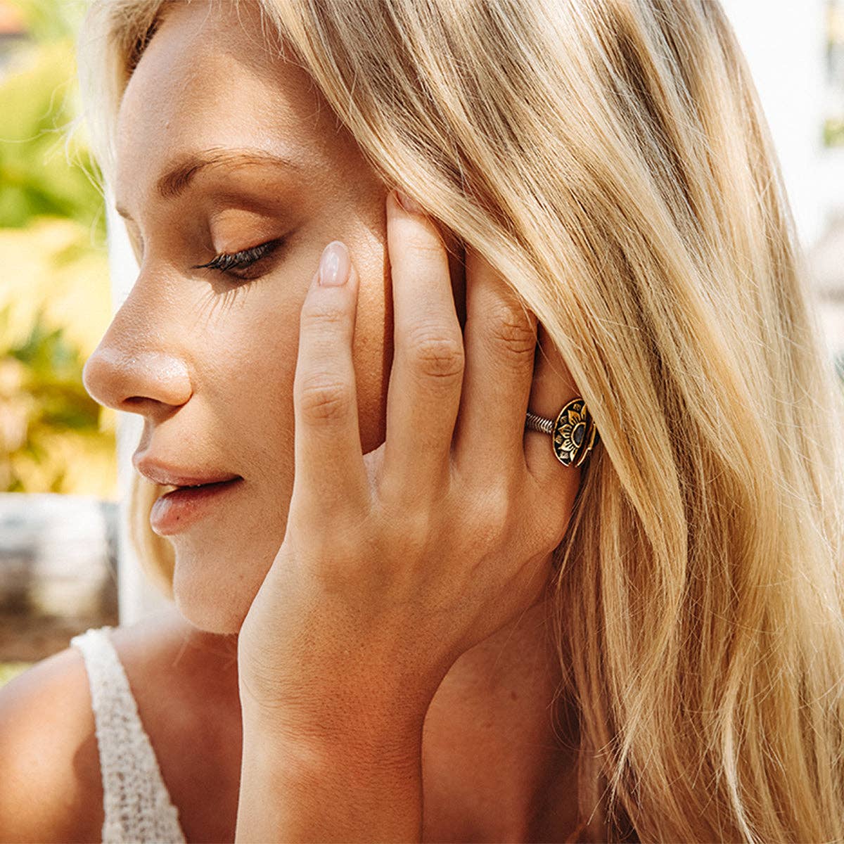 A woman with long blonde hair, eyes closed, gently touches her face while wearing a sleeveless top and the bahgsu jewels Natural Mystic Ring - Moonstone. The sterling silver ring features an ornate leaf design; greenery softly blurs in the background.