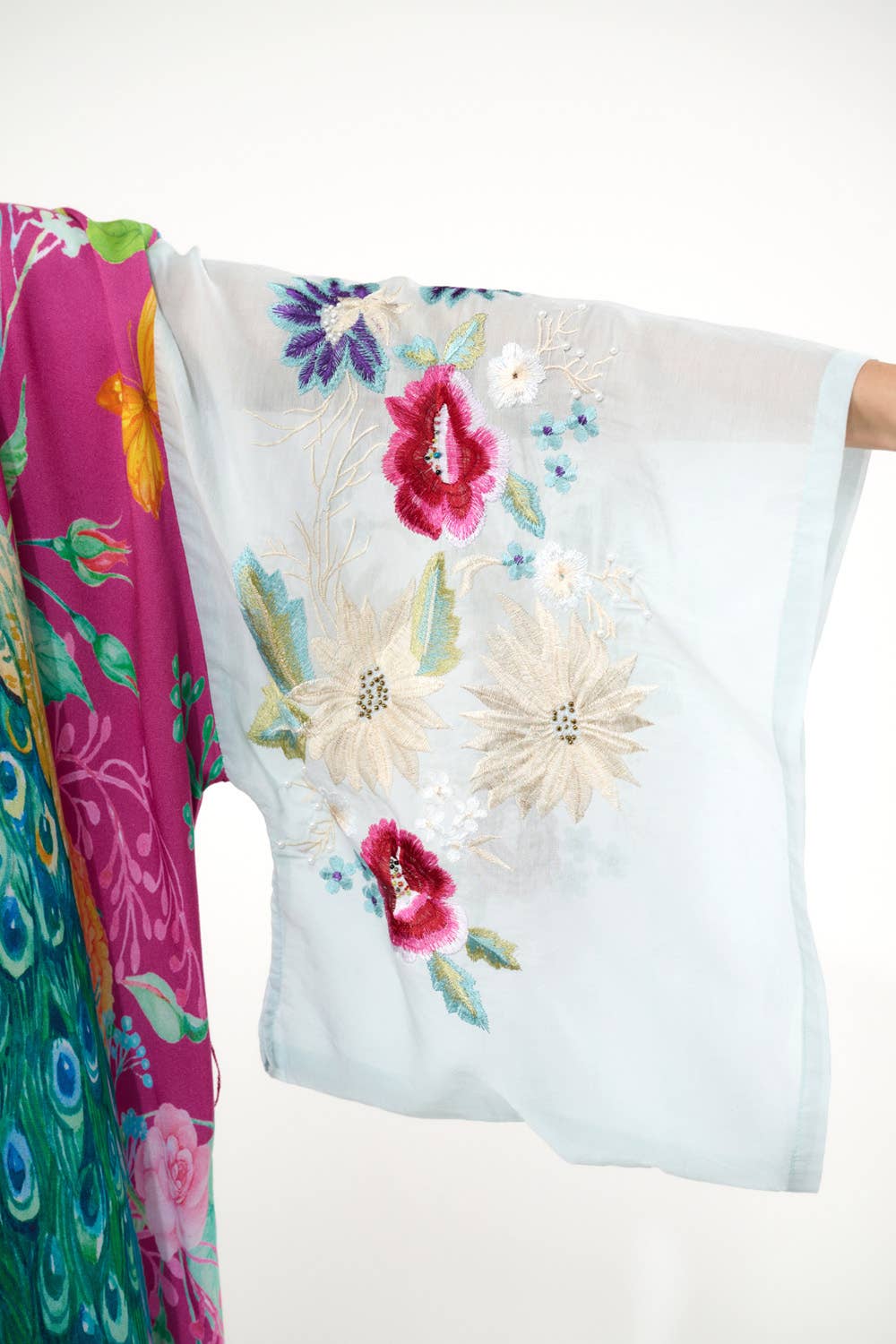 A close-up of the Aratta Marie Antoinette Kimono shows wide, light blue sleeves embroidered with white and magenta flowers, and a vibrant main body featuring a peacock and floral motif—a unique statement piece.
