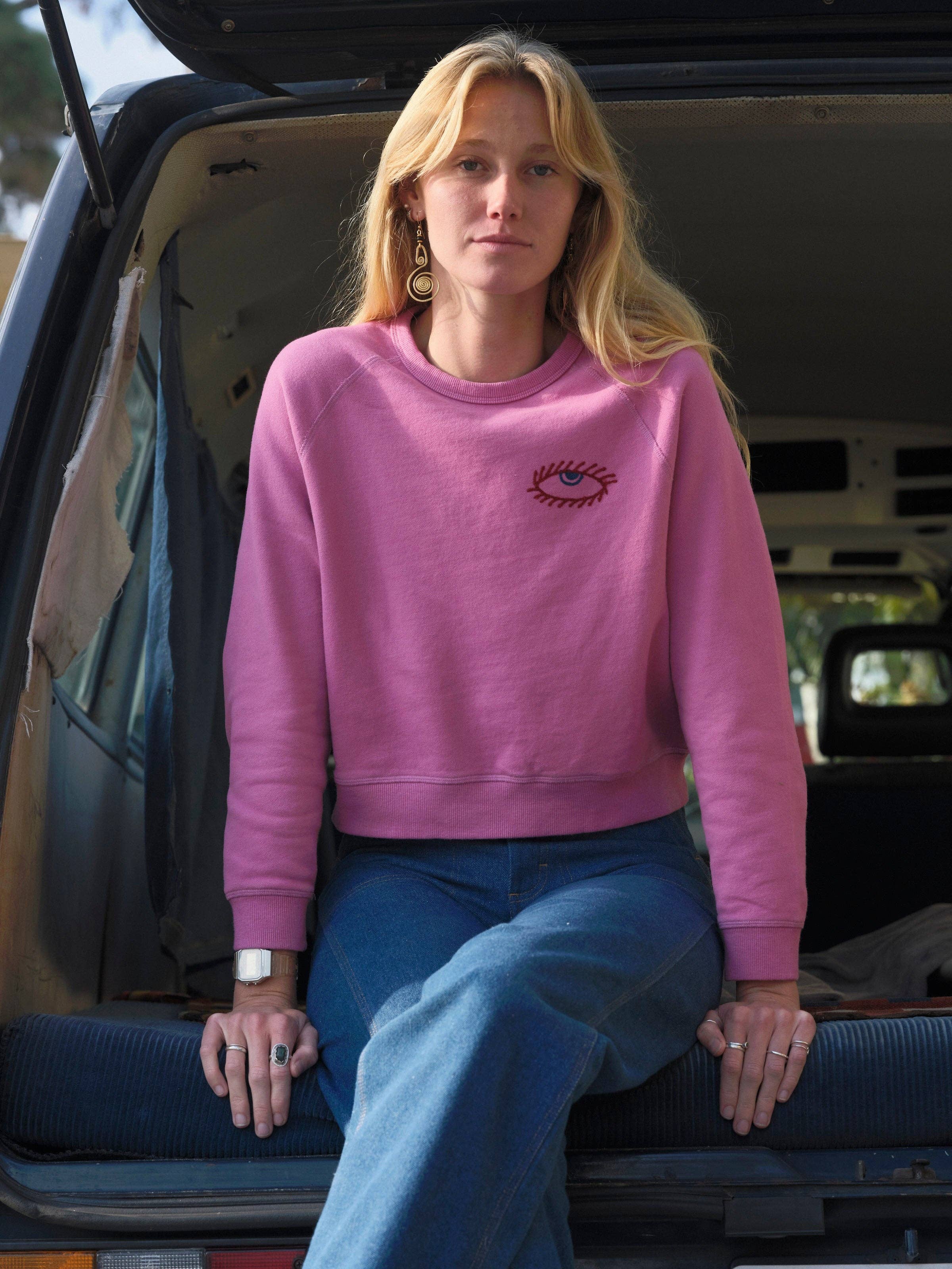 A woman with long blonde hair wearing the Mollusk Visions Crew sweatshirt and blue jeans sits in the back of a van with the door open, gazing calmly at the camera.