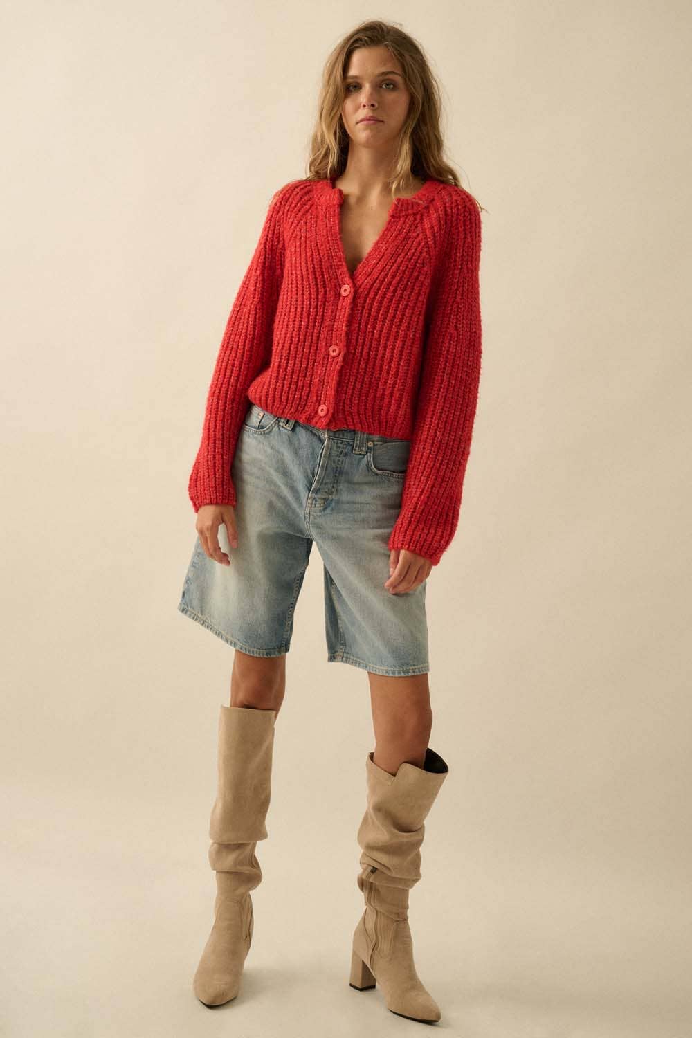 A woman stands facing forward against a beige background, wearing the Promesa USA Confetti Button-Front Cardigan Sweater in red, light-wash knee-length denim shorts, and beige slouchy heeled boots.