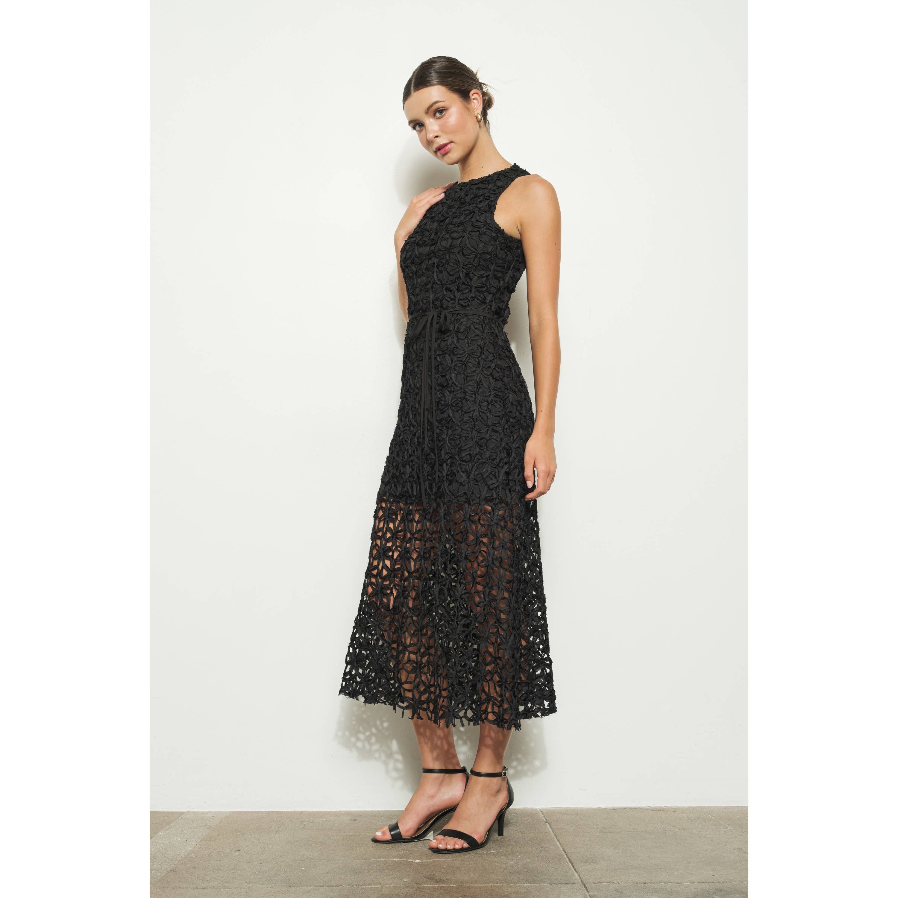 A woman stands against a white wall, wearing the INA Sleeveless Crochet Lace Overlay Midi Dress with a high neckline and sheer midi skirt. Her hair is tied back and she pairs the look with black strappy heels.