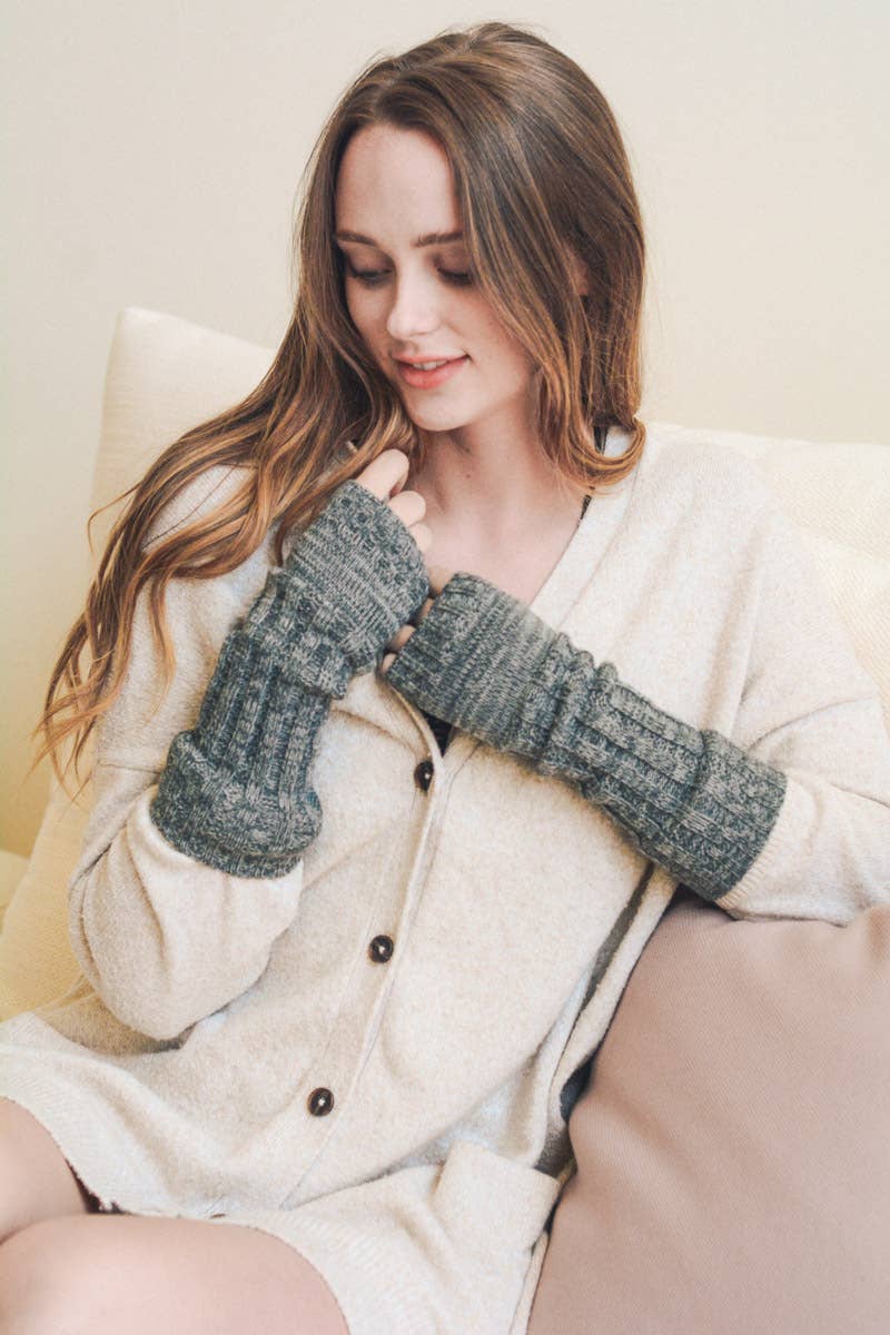 A woman with long brown hair sits on a beige couch, smiling as she looks down at her cozy Ribbed Arm Warmers from Leto Accessories, paired with a cream button-up cardigan.