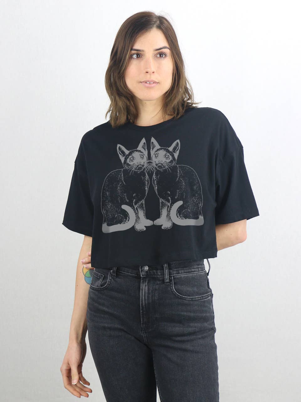 A woman with shoulder-length brown hair wears the Supermaggie @ Faire Kitties Clover Crop Tee, a black cropped cotton tee featuring two black cats, styled with high-waisted black jeans against a plain white background.