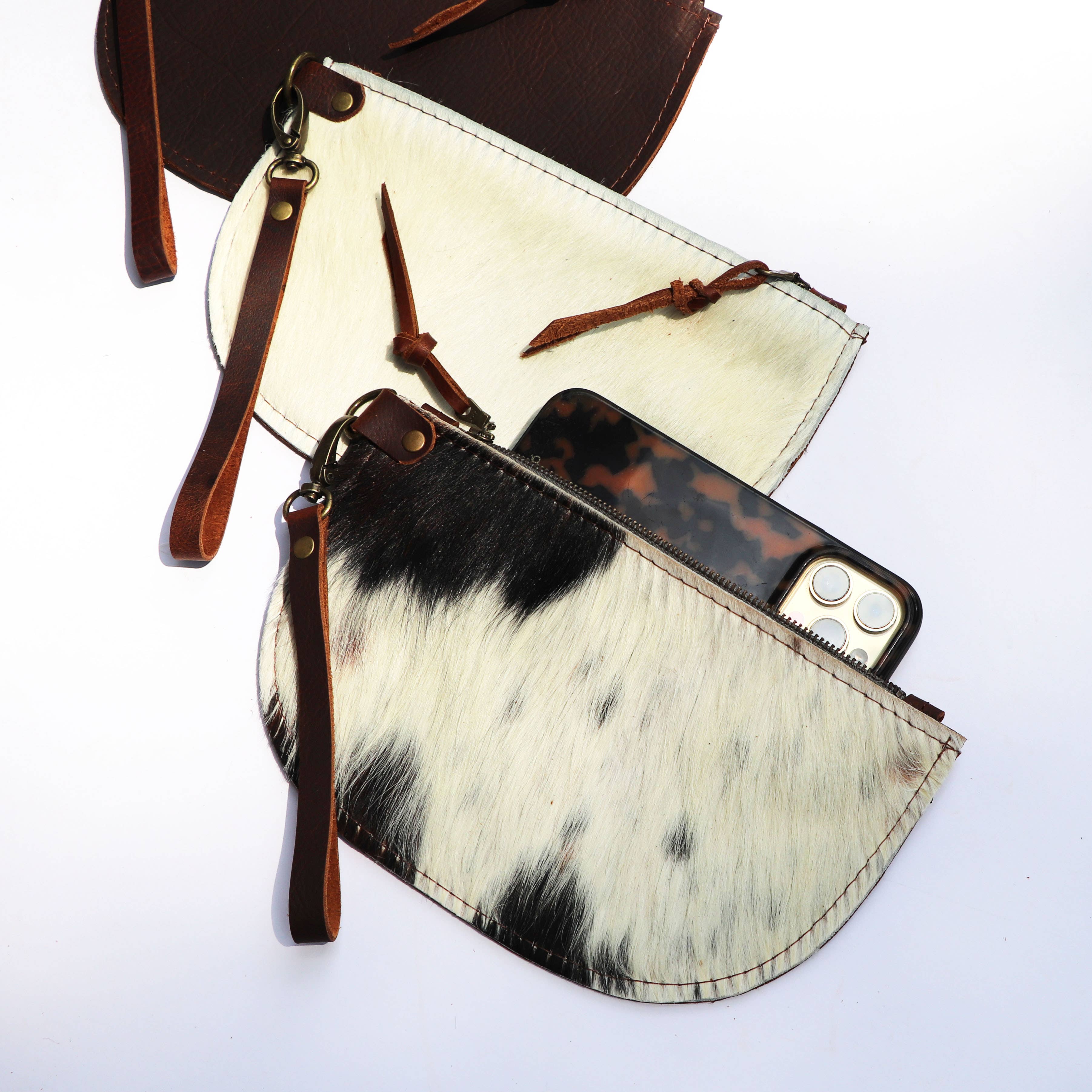 Two Hawks & Doves Large Half Moon Cowhide and Leather Zip Pouches with detachable wristlets are displayed—one open showing a phone and sunglasses. Features: spacious half-moon design, premium cowhide leather, versatile carry options.