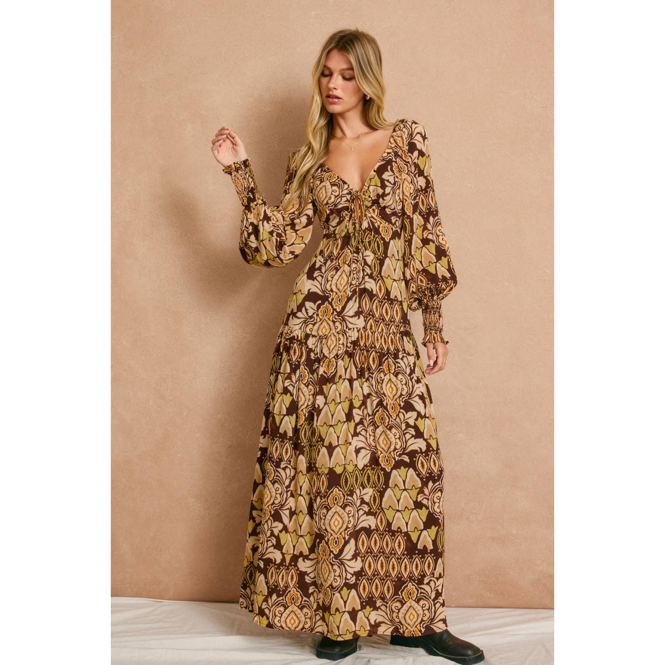 A woman wears the Stone Ember Keyhole Long Sleeve Maxi Dress by Dress Forum, featuring earthy tones and a deep neckline, standing against a beige backdrop.