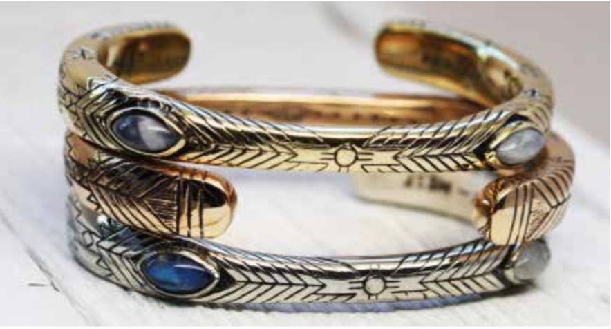Two intricately engraved bahgsu jewels Medicine Wheel Cuffs—one gold, one Indian silver—each featuring an oval moonstone or turquoise inlay and arrow motifs, are stacked together on a white surface.