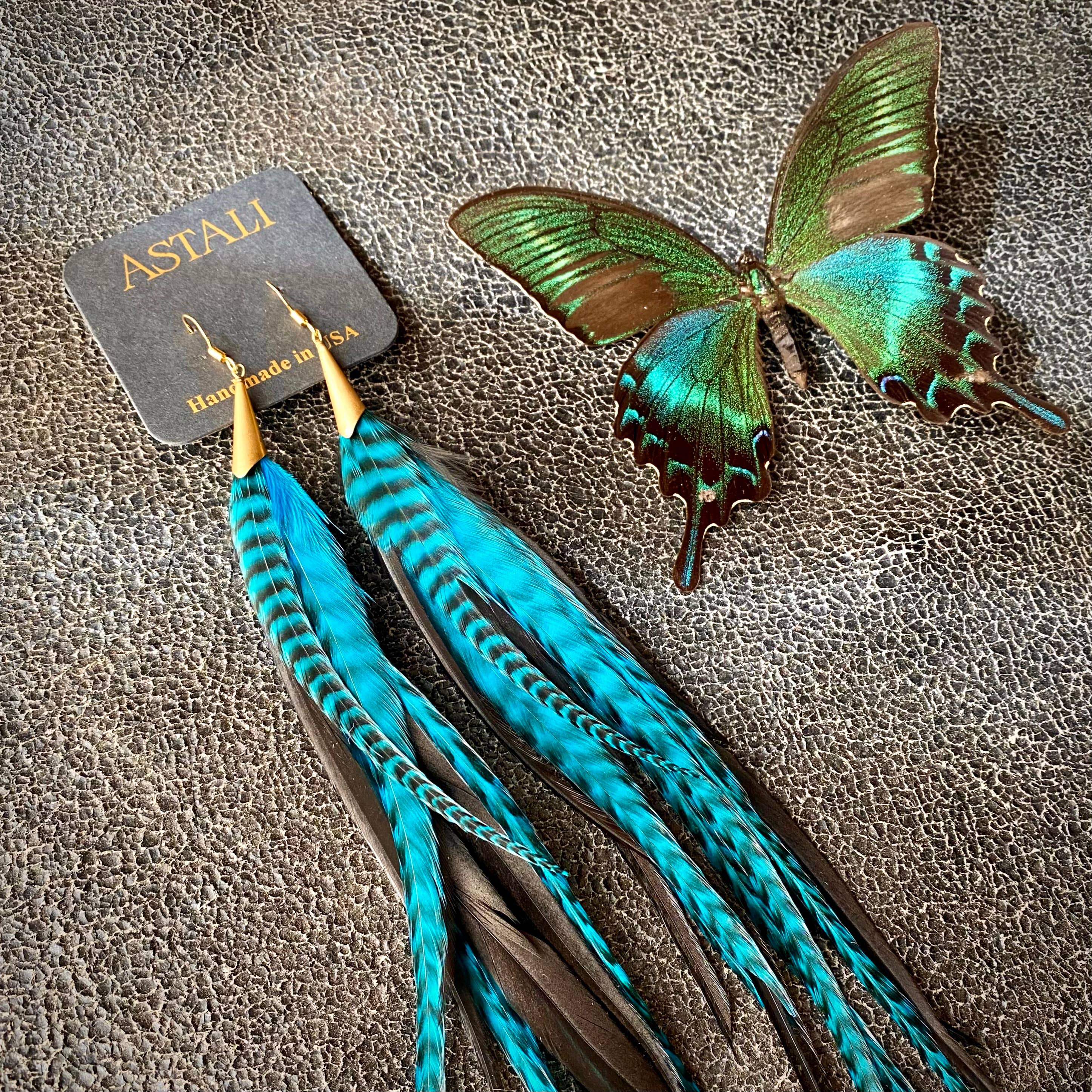 A pair of ASTALI Long Feather & Cone Earrings in Turquoise Grizzly & Black/Gold rests beside a green and brown butterfly brooch on a textured brown surface.