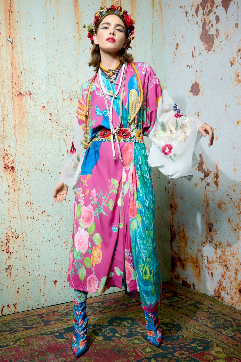 A woman poses in the Marie Antoinette Kimono by Aratta, featuring vibrant pink, blue, and yellow floral patterns with wide embroidered sleeves and a matching flower crown, standing on a patterned rug before a distressed wall.