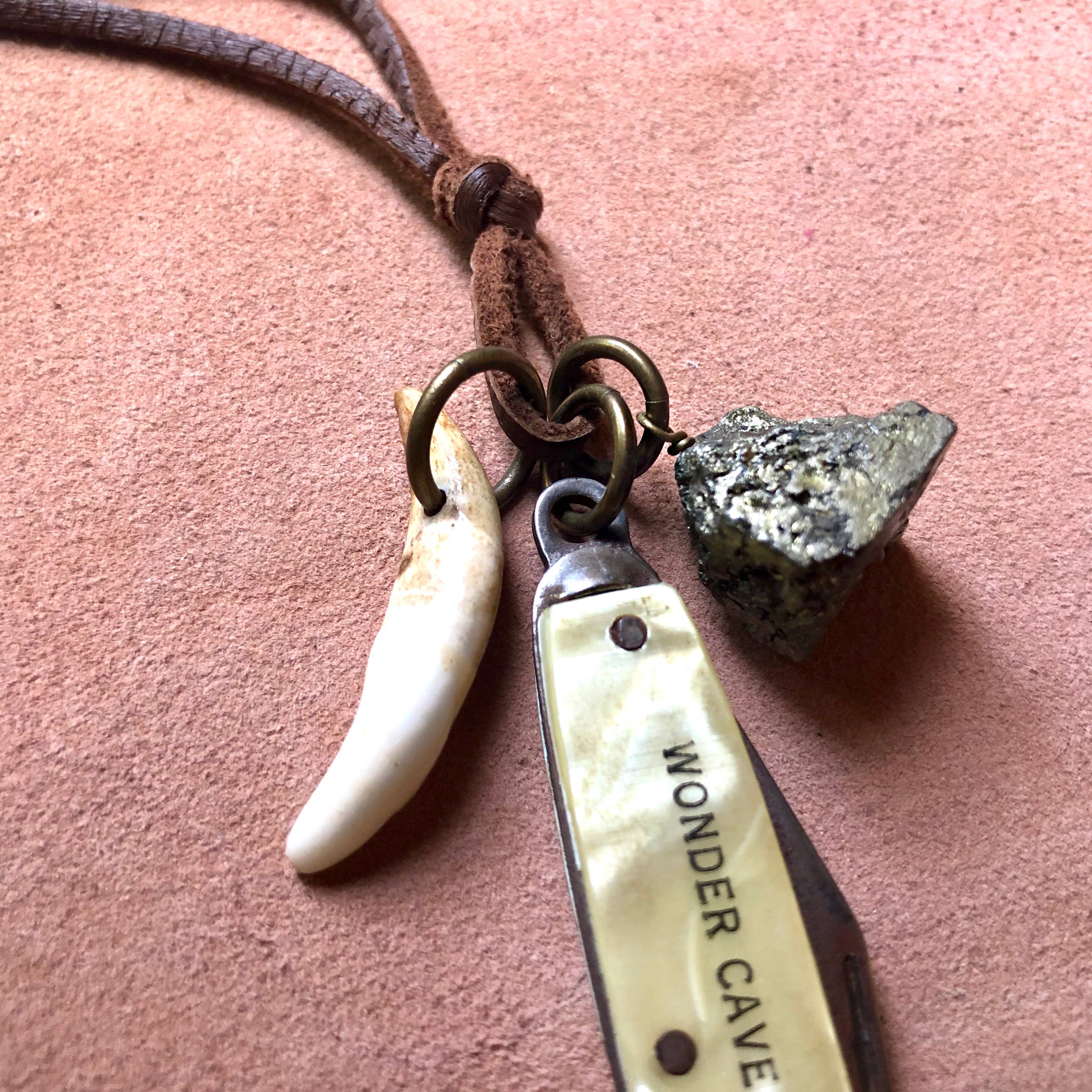 The ASTALI Vintage Pocket Knife & Coyote Tooth Necklace features a white coyote tooth, WONDER CAVE pocket knife pendant, and rough pyrite nugget on deerskin leather, shown on a pinkish-brown surface.