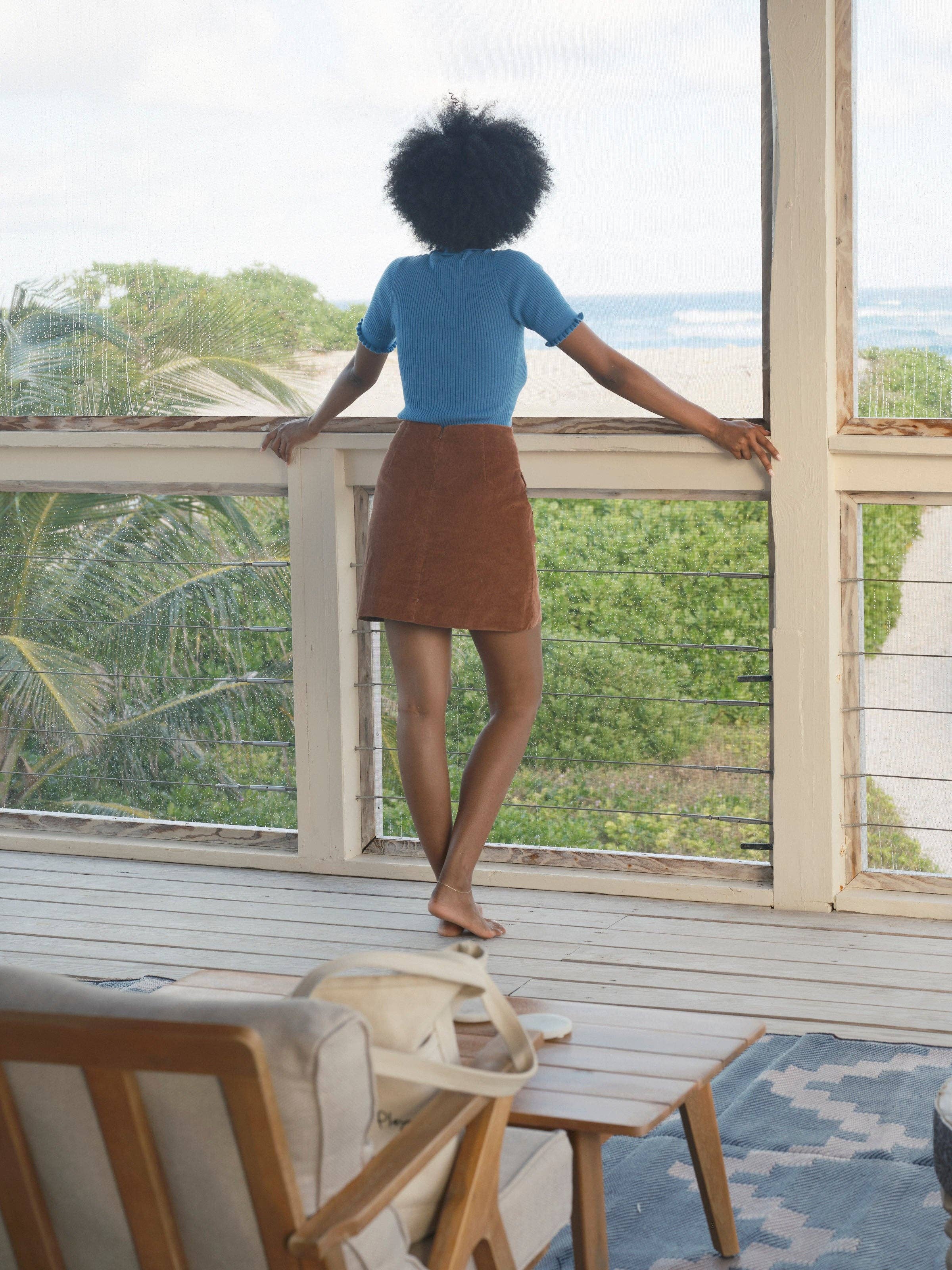 A woman with curly hair stands barefoot on a porch, overlooking greenery and water. She wears the Mollusk Belle Tee, a vintage-inspired blue top with a Peter Pan collar, paired with a brown skirt, hands resting on the railing.