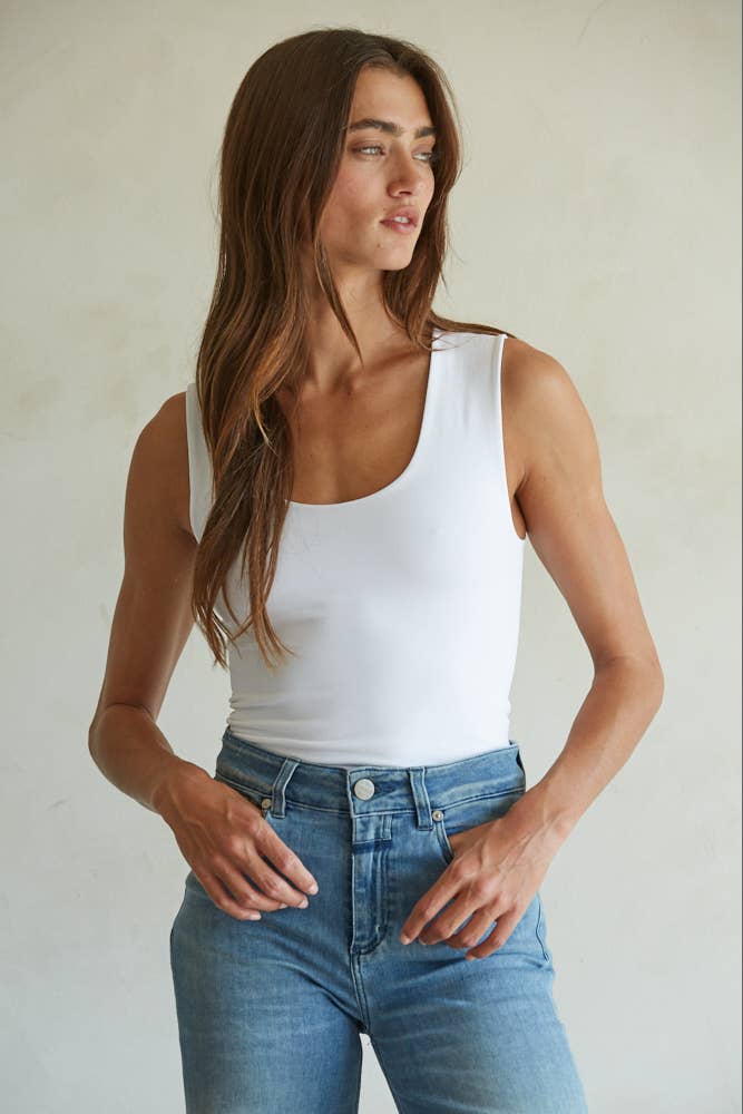 A woman with long brown hair wears a white By Together Double Layered Seamless Top and blue jeans, standing against a light background and looking to her left with a relaxed expression.