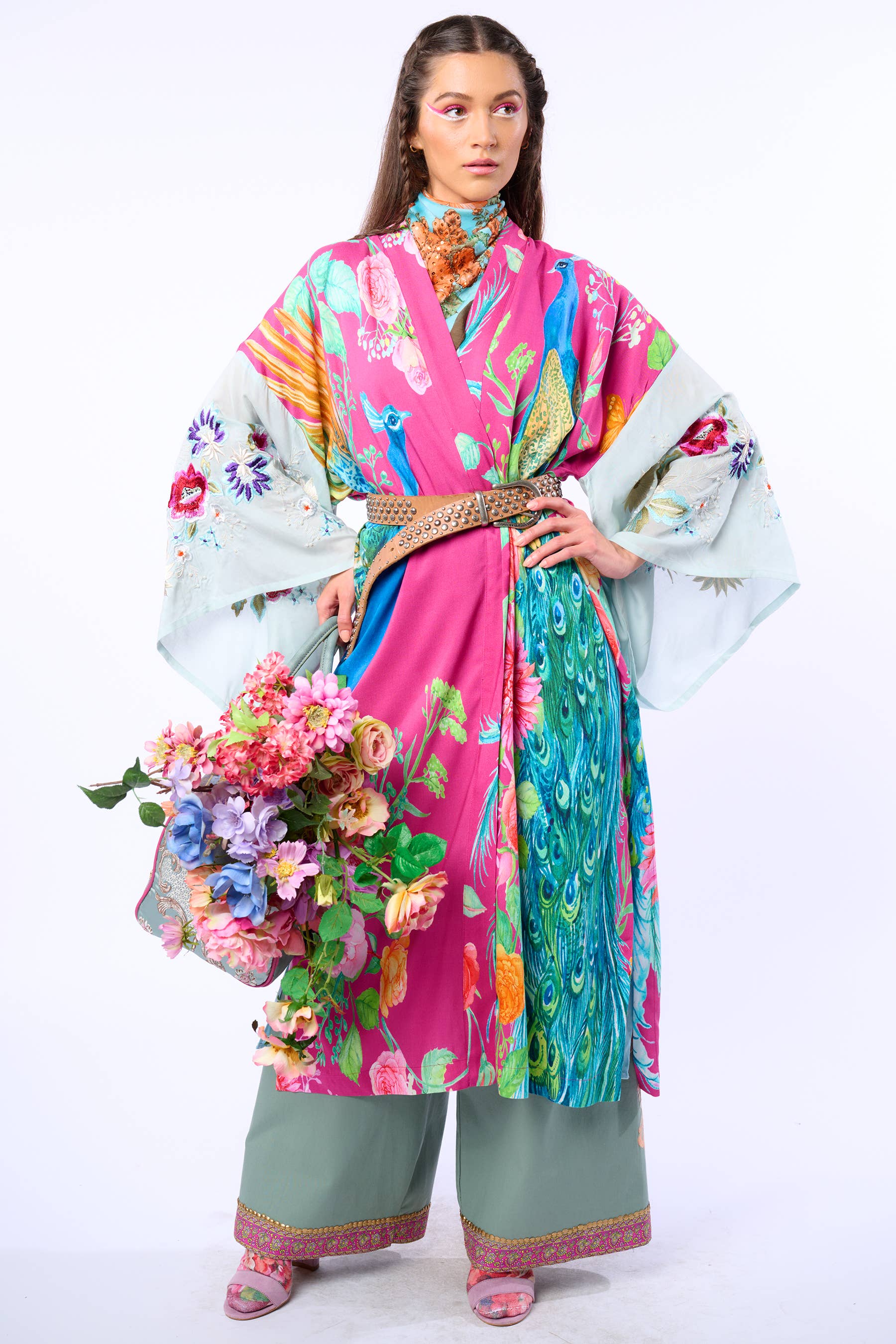 A woman poses holding a basket of flowers in the Marie Antoinette Kimono by Aratta, featuring embroidered floral sleeves, wide green pants, and pink sandals—her wavy brown hair completes this bohemian statement look.