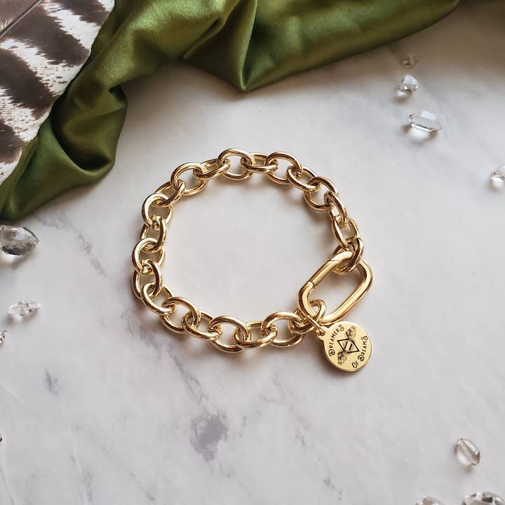 The Chunky Cable Chain Gold Plated Bracelet by Dreamers of Dreams features a round engraved charm and rectangular clasp, displayed on marble with green fabric and decorative crystals.