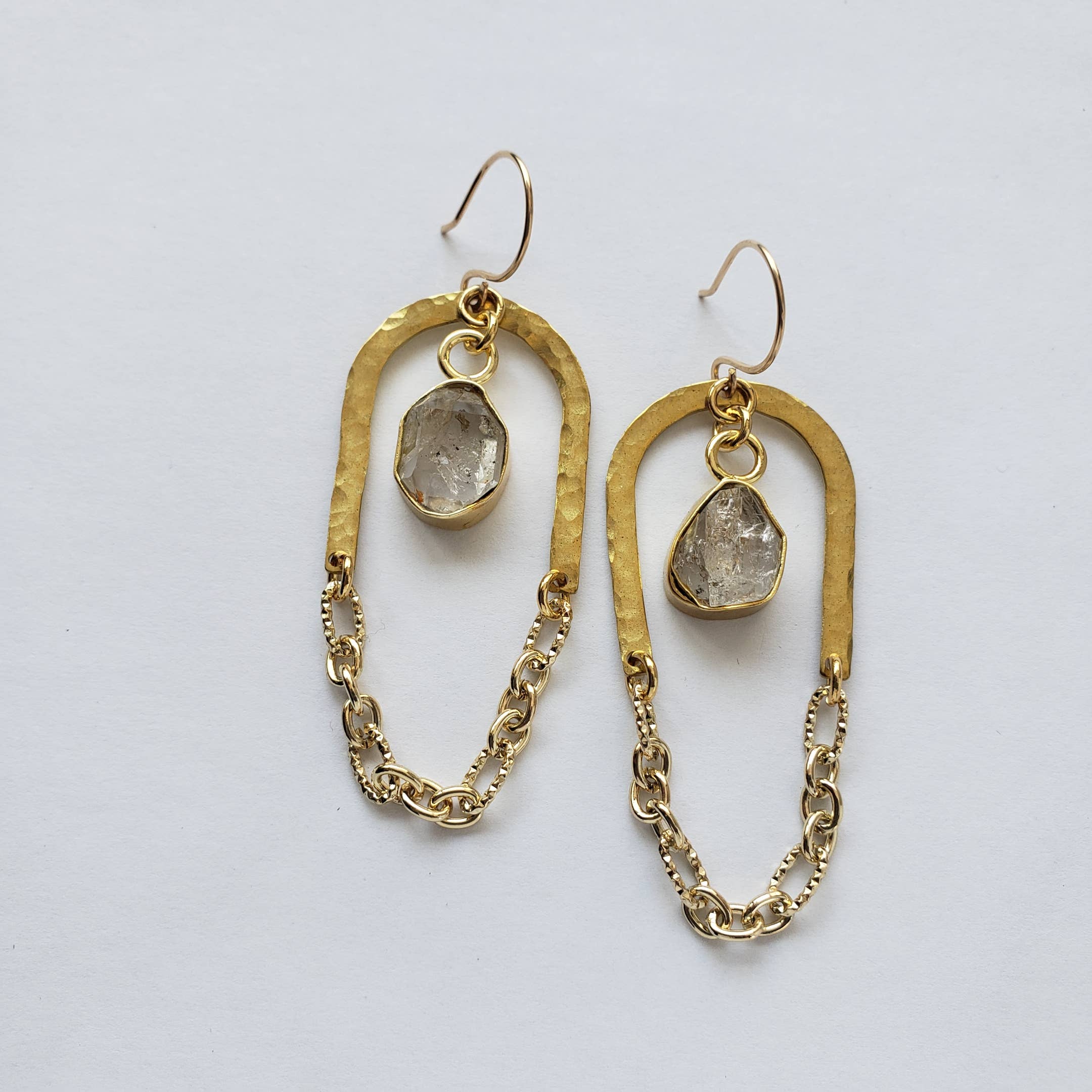 The Gold + Glam Arch Chain Earrings by Dreamers of Dreams feature irregular gold oval hoops with rough-cut clear stones and decorative gold chains at the bottom, shown on a white background.