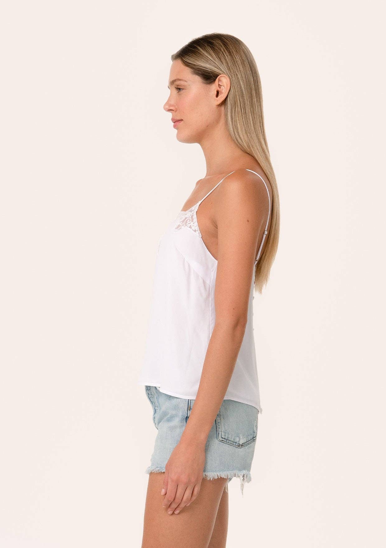 Woman wearing a white Button Back Embroidered Lace Cami with delicate straps paired with light denim shorts in profile view