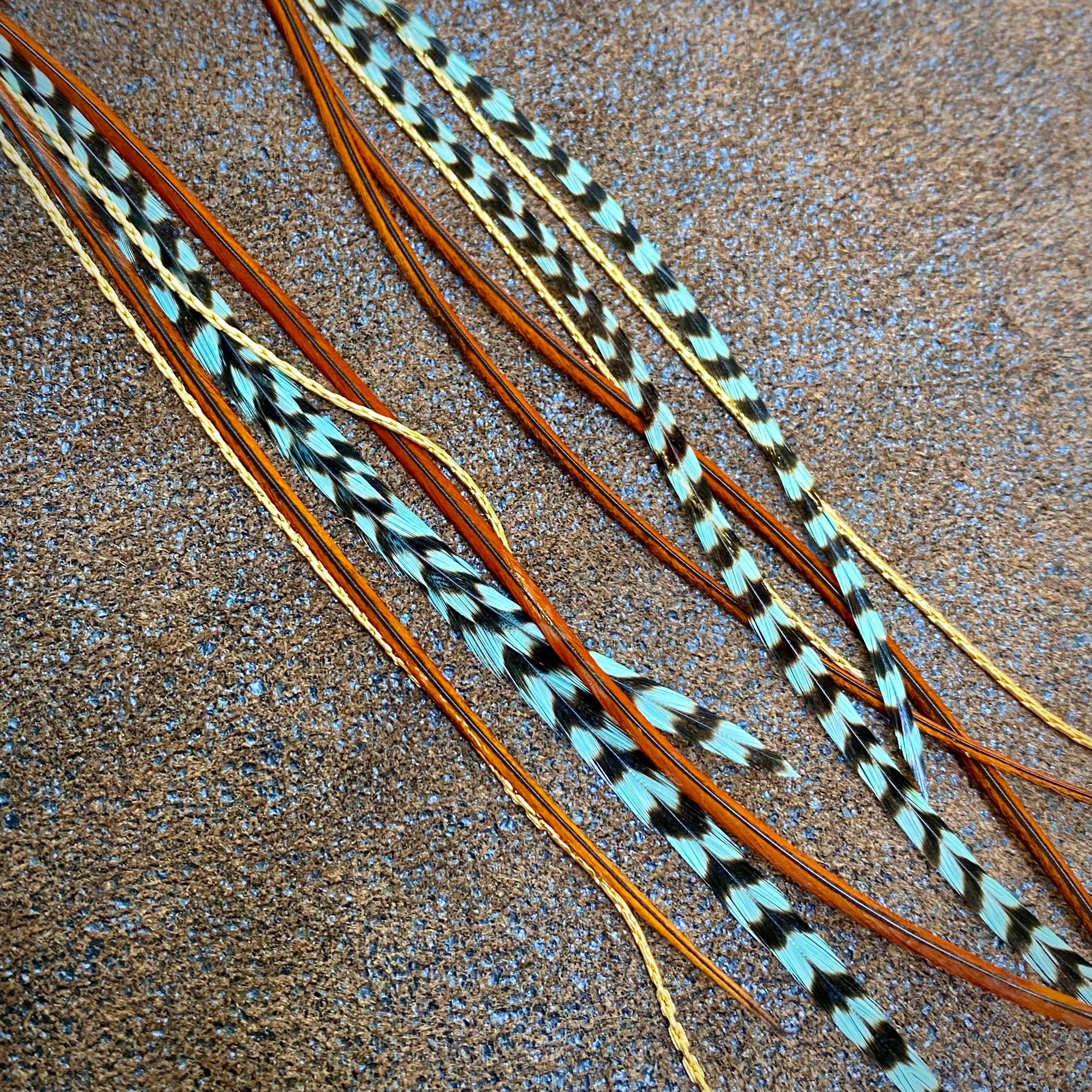 Long, slender feathers with black and teal grizzly stripes, plus fawn and gold accents, are displayed on a brown and blue-speckled surface—capturing the look of ASTALI’s Turquoise & Mini Feather Earrings - Teal Grizzly & Fawn/Gold.
