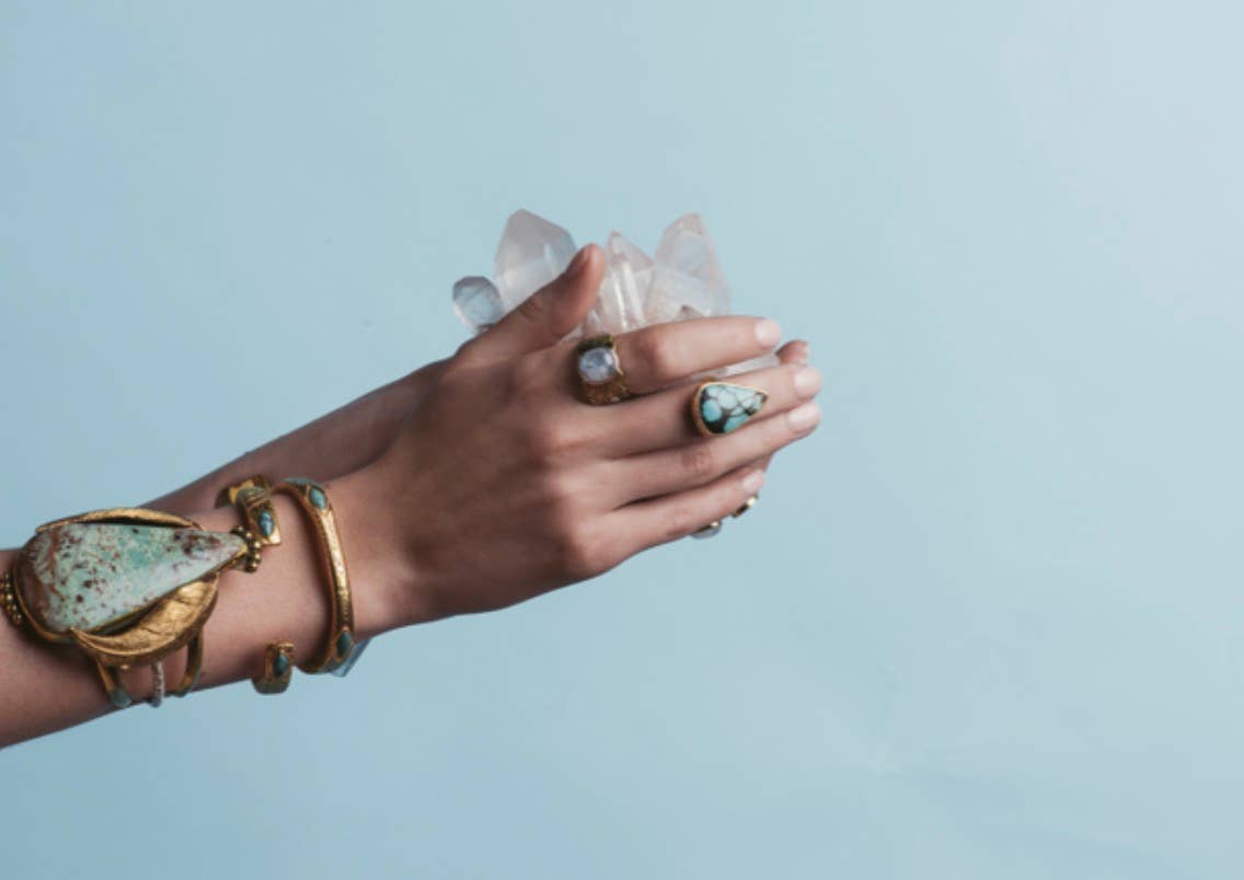 A hand wearing the bahgsu jewels Scarab Ring in gold and turquoise holds clear quartz crystals against a light blue background.