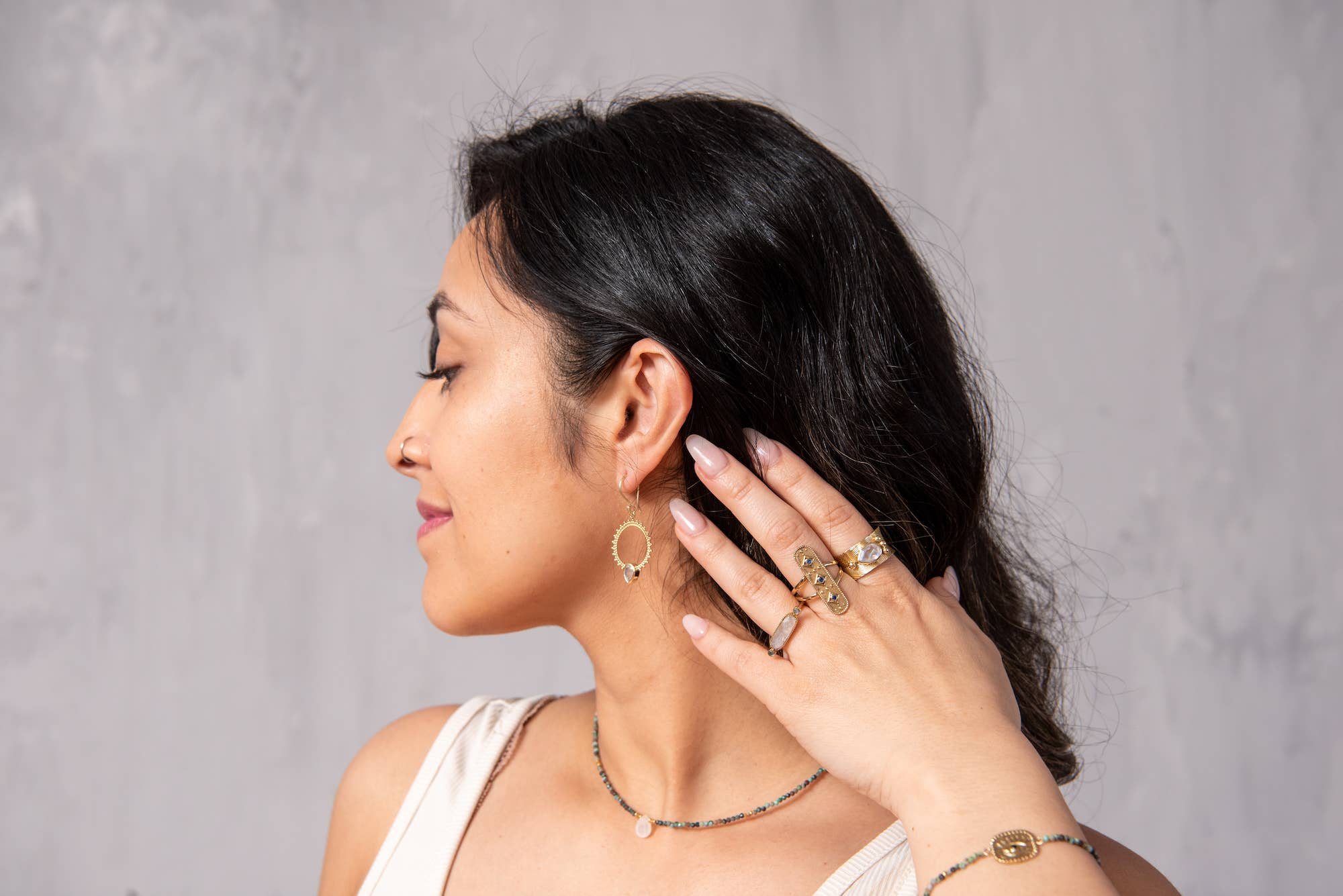 A woman with long dark hair looks to the side, showcasing gold earrings, a necklace, and bracelet. She highlights her manicured nails and wears Baizaar’s Sapphire Healer Ring with a sleeveless light-colored top.