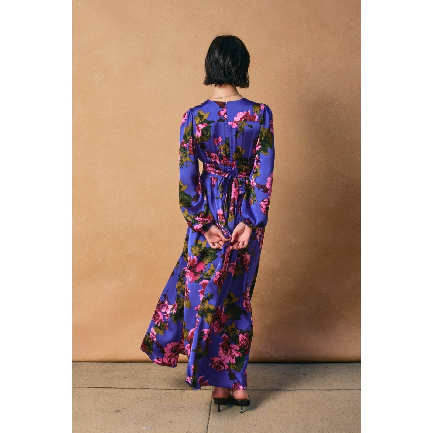A woman with short dark hair stands with her back to the camera, modeling the Mesmerizing Essence Pleated Waist Maxi Dress by Dress Forum—a floral satin maxi dress with vibrant pink patterns, a pleated waist, and side slit, set against a tan backdrop.