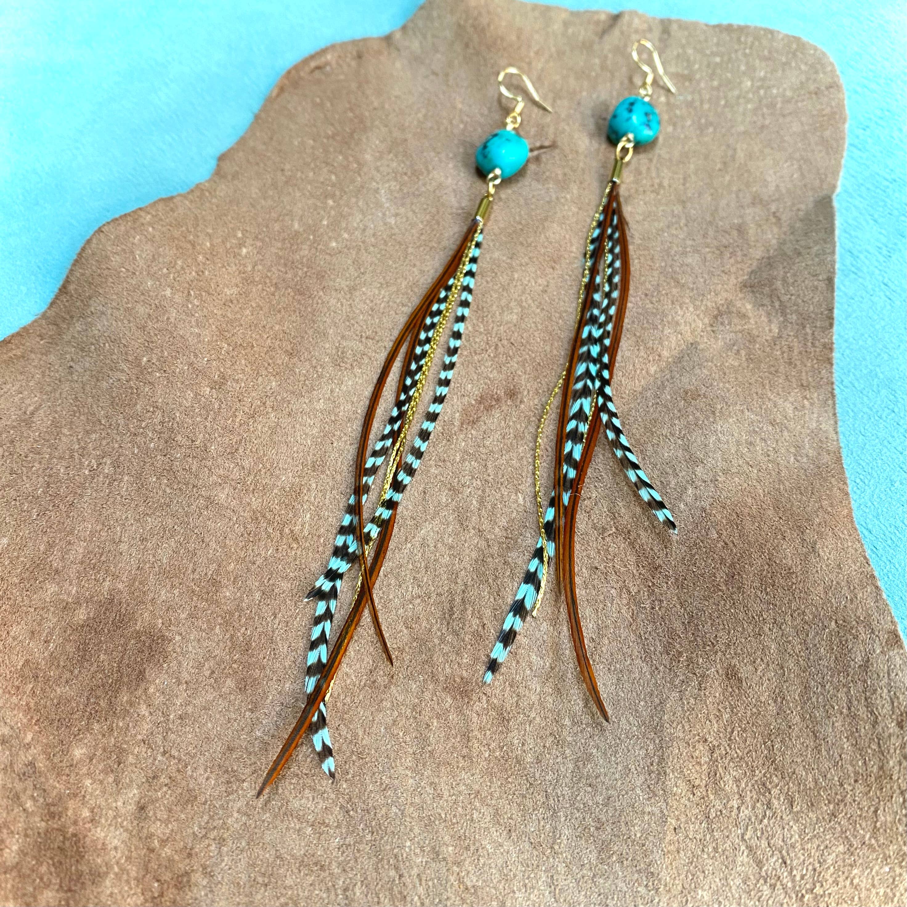 ASTALI Turquoise & Mini Feather Earrings feature teal grizzly and fawn/gold feathers with long, dangling beads, showcased on a brown textured surface against a blue background.