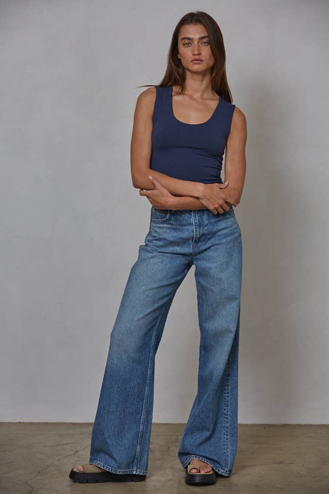 A woman stands against a plain gray wall, wearing By Together’s Double Layered Seamless Top in navy blue, wide-leg blue jeans, and black platform sandals. Her arms are loosely crossed as she gazes forward with a neutral expression.
