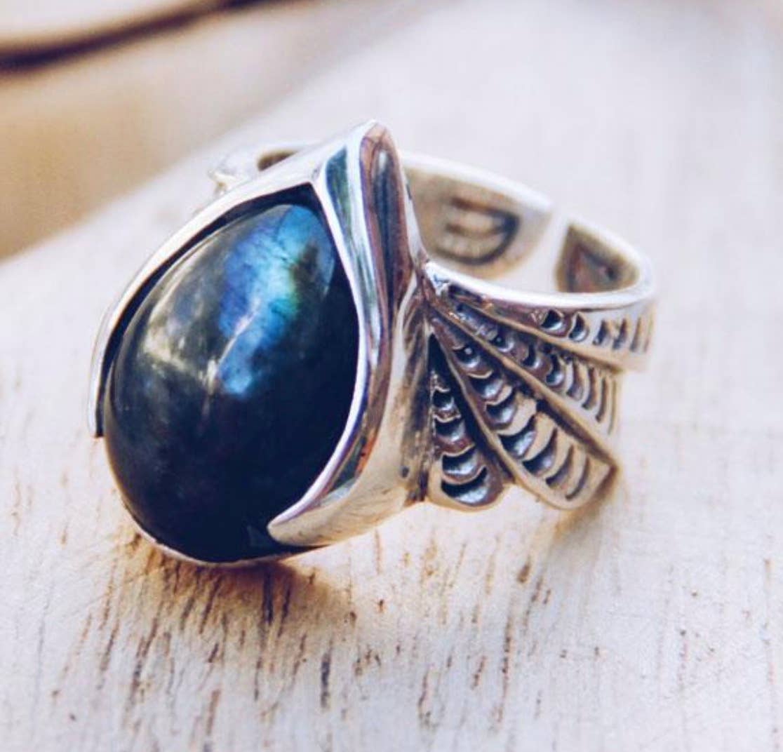 The bahgsu jewels Winged Ring showcases a silver design with wing motifs and an iridescent oval gemstone, set in an ornate style and displayed on a light wooden surface.