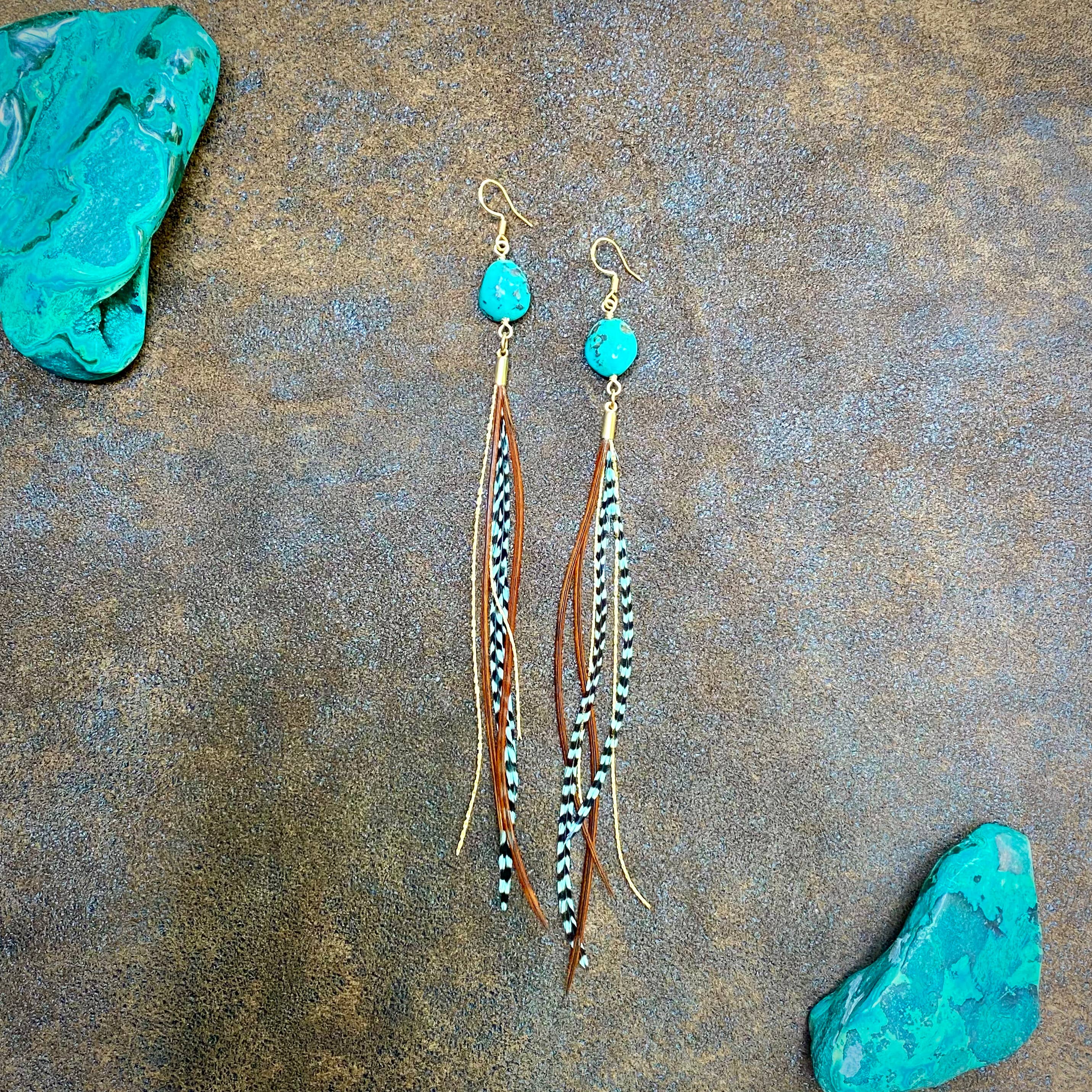 ASTALIs Turquoise & Mini Feather Earrings – Teal Grizzly & Fawn/Gold feature boho-style turquoise stones, leather tassels, and striped feathers. Shown on a textured brown surface with turquoise rocks surrounding the earrings.