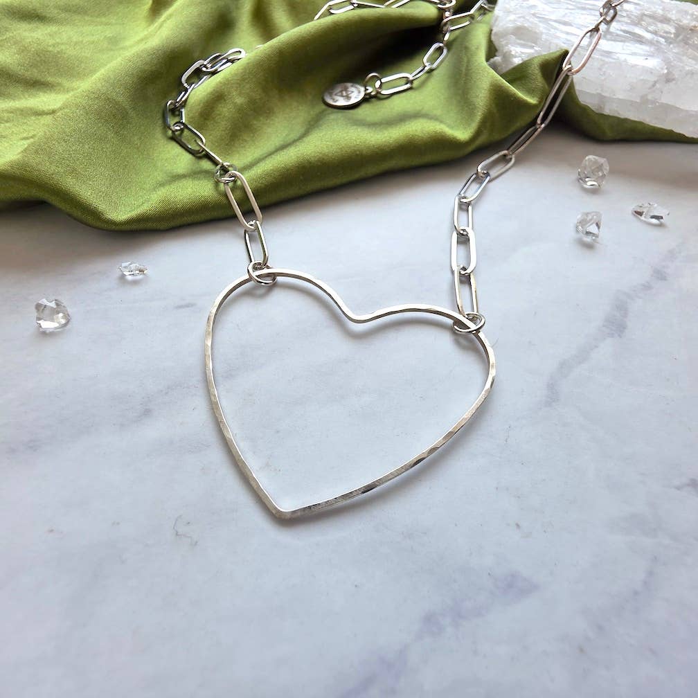 The Dreamers of Dreams Big Silver Heart - Paperclip Chain Necklace, featuring a large hammered heart charm on a silver-plated brass chain, is displayed on marble with green fabric, crystals, and decorative stones.