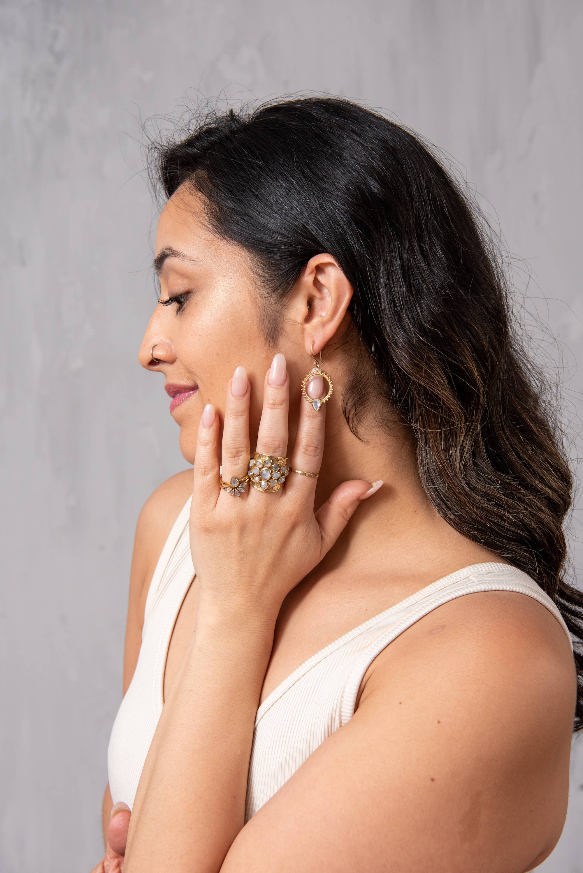 A woman with dark wavy hair poses in profile, showcasing Baizaar's Brass Nesting Moonstone Ring Set. She wears a sleeveless cream top and gently touches her face, highlighting the artisan rings against a plain grey background.