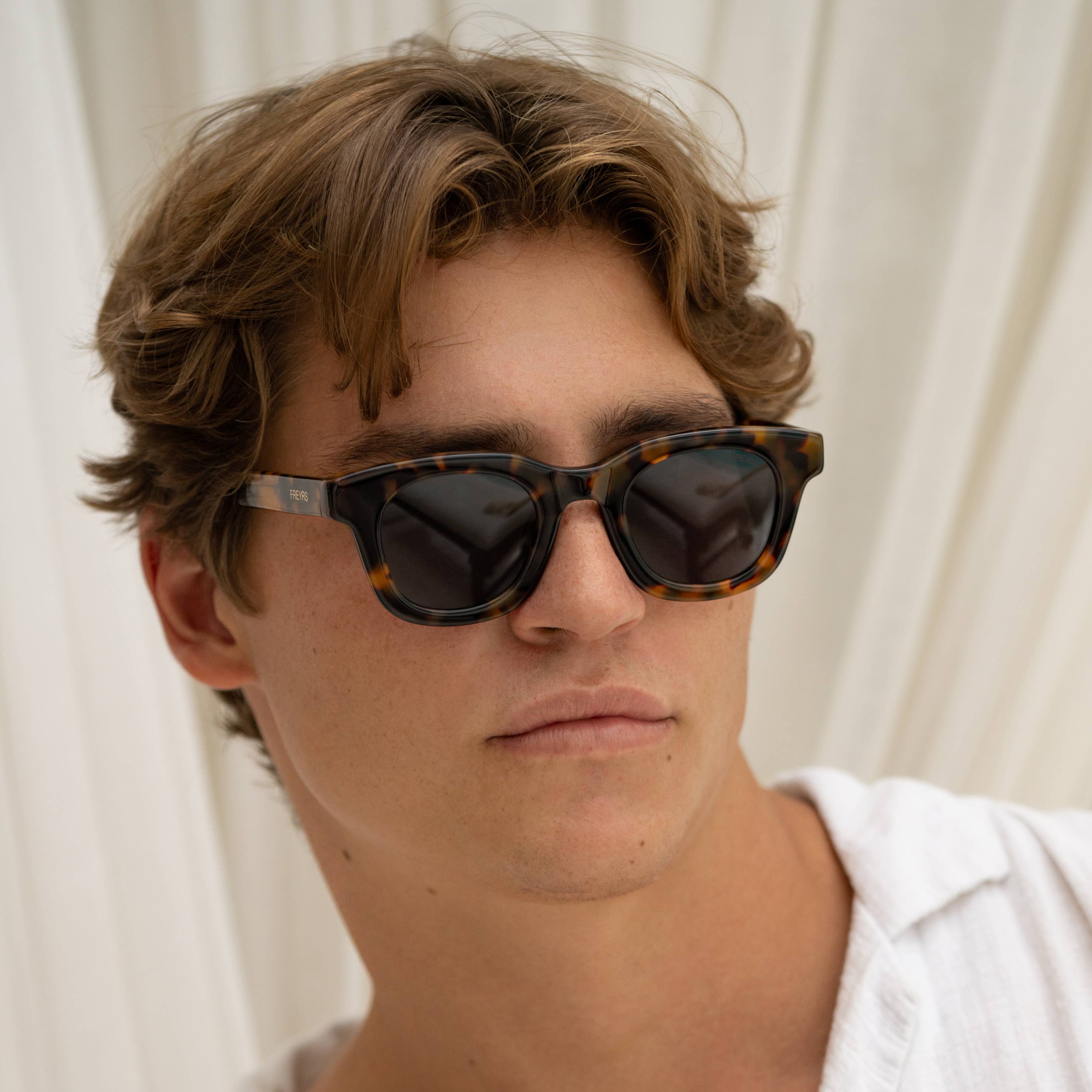 A young man with wavy brown hair wears FREYRS Eyewear Deni Acetate Round Mens Sunglasses with UVA/UVB protection and a white shirt, standing confidently before a light, draped backdrop.