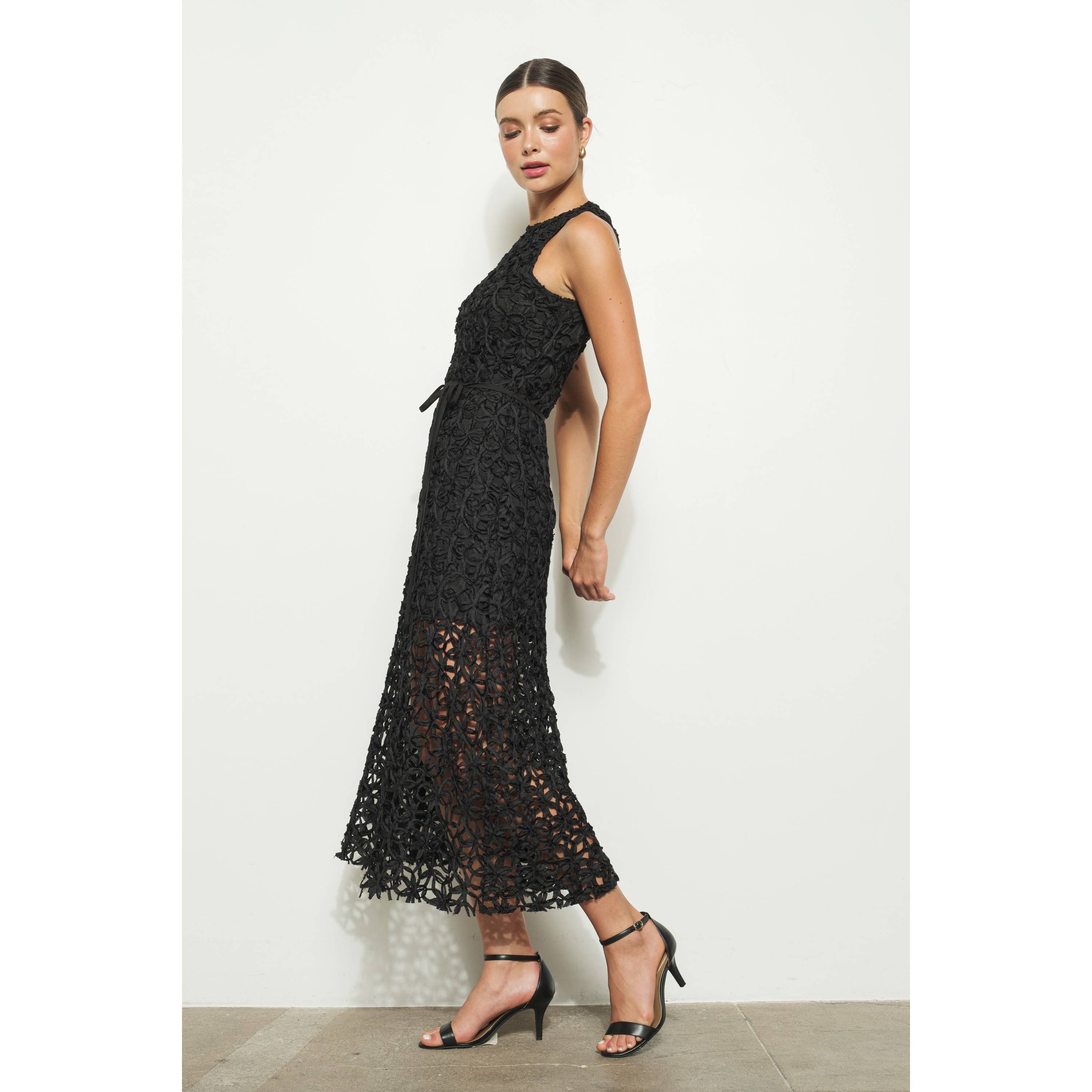 A woman poses against a white wall wearing the INA Sleeveless Crochet Lace Overlay Midi Dress with a sheer skirt, paired with black strappy high-heeled sandals.