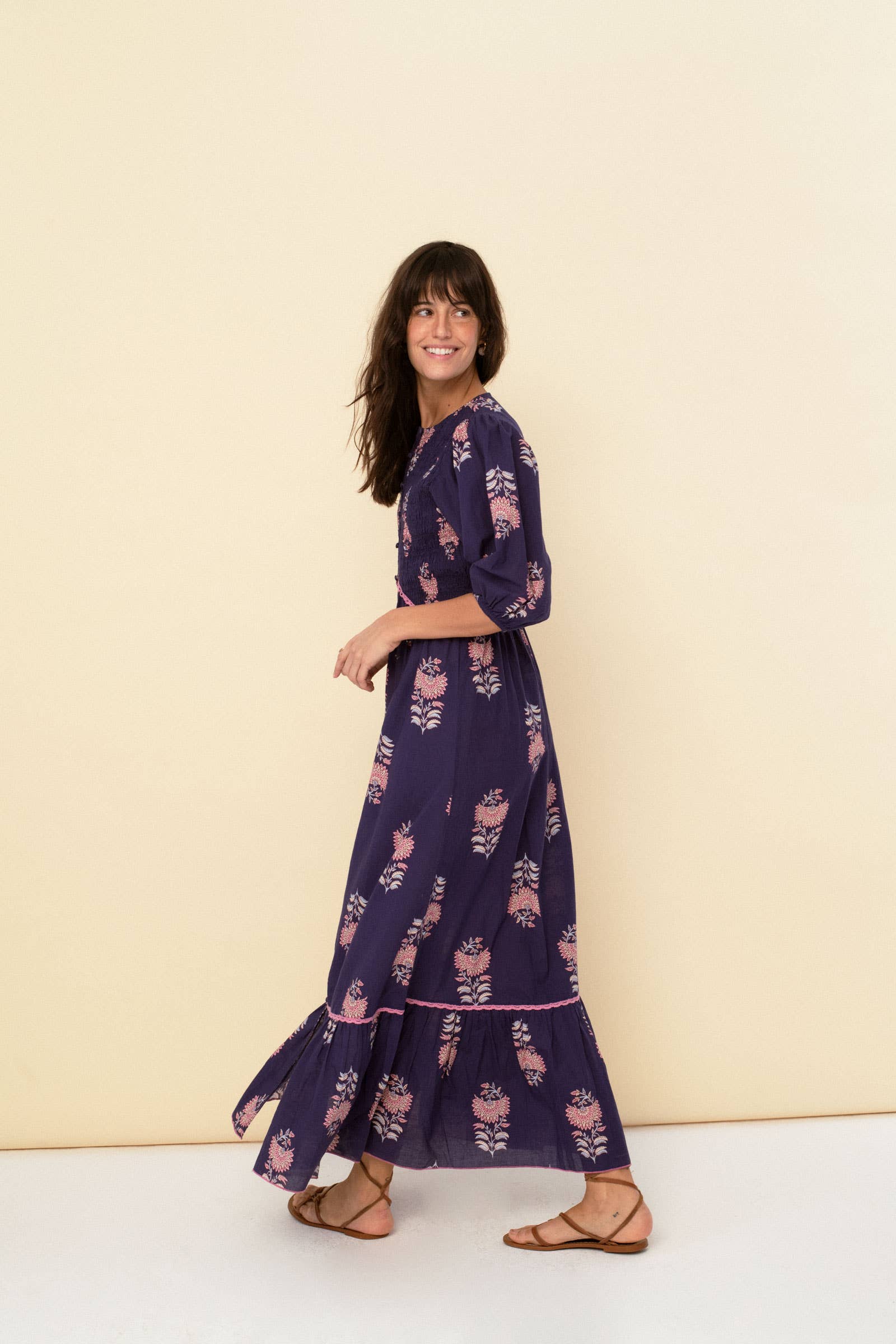 A woman with long brown hair smiles in the Magnolia dress blue by Sonmer Shop USA—an Edwardian-inspired, navy floral ankle-length maxi with elbow sleeves—styled with brown sandals, standing before a plain beige background.