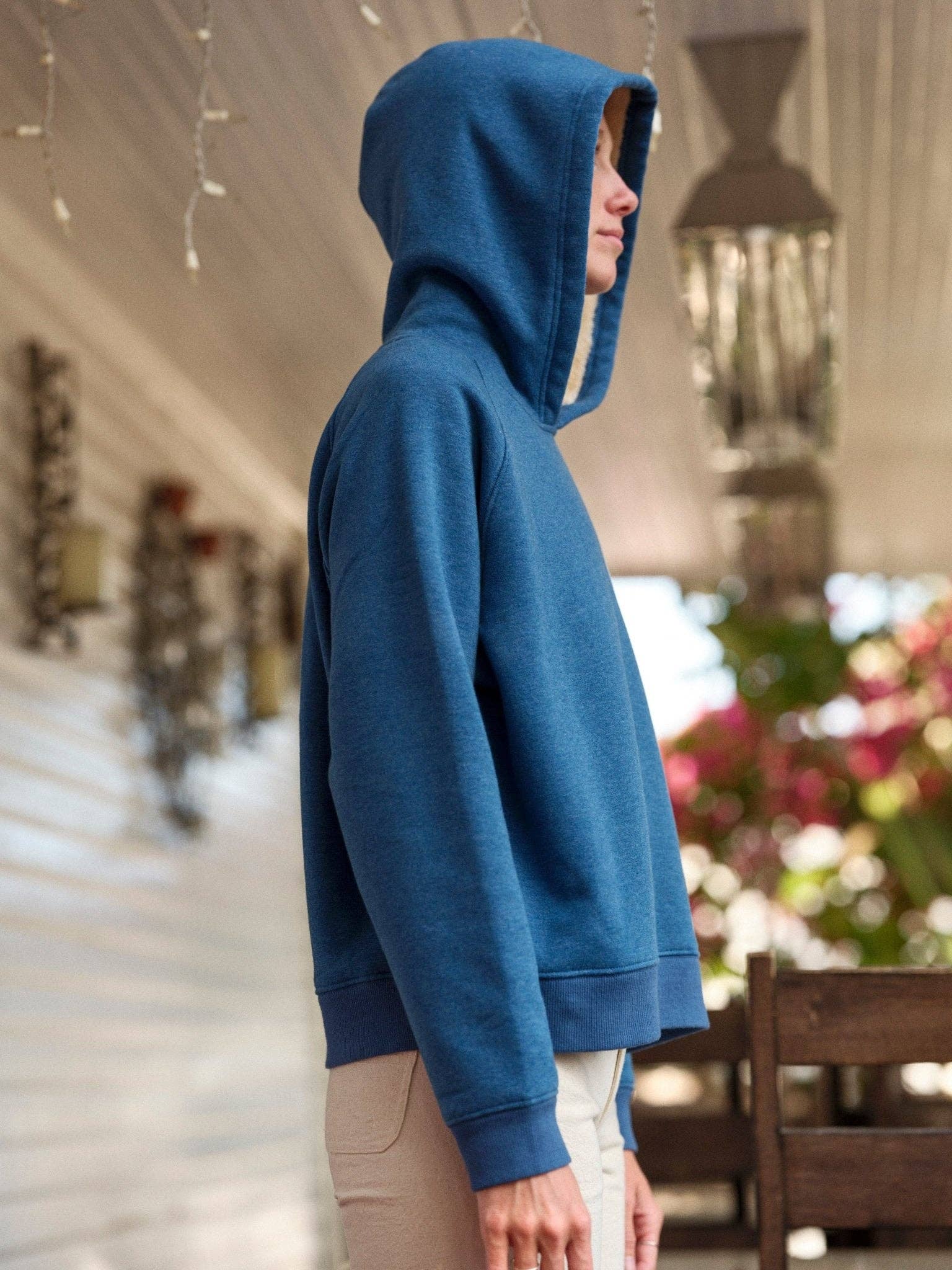 A person stands in profile on a porch under string lights, wearing Mollusk’s Cozy Sherpa-Lined Sweatshirt in blue with beige pants; plants and furniture appear blurred in the background.