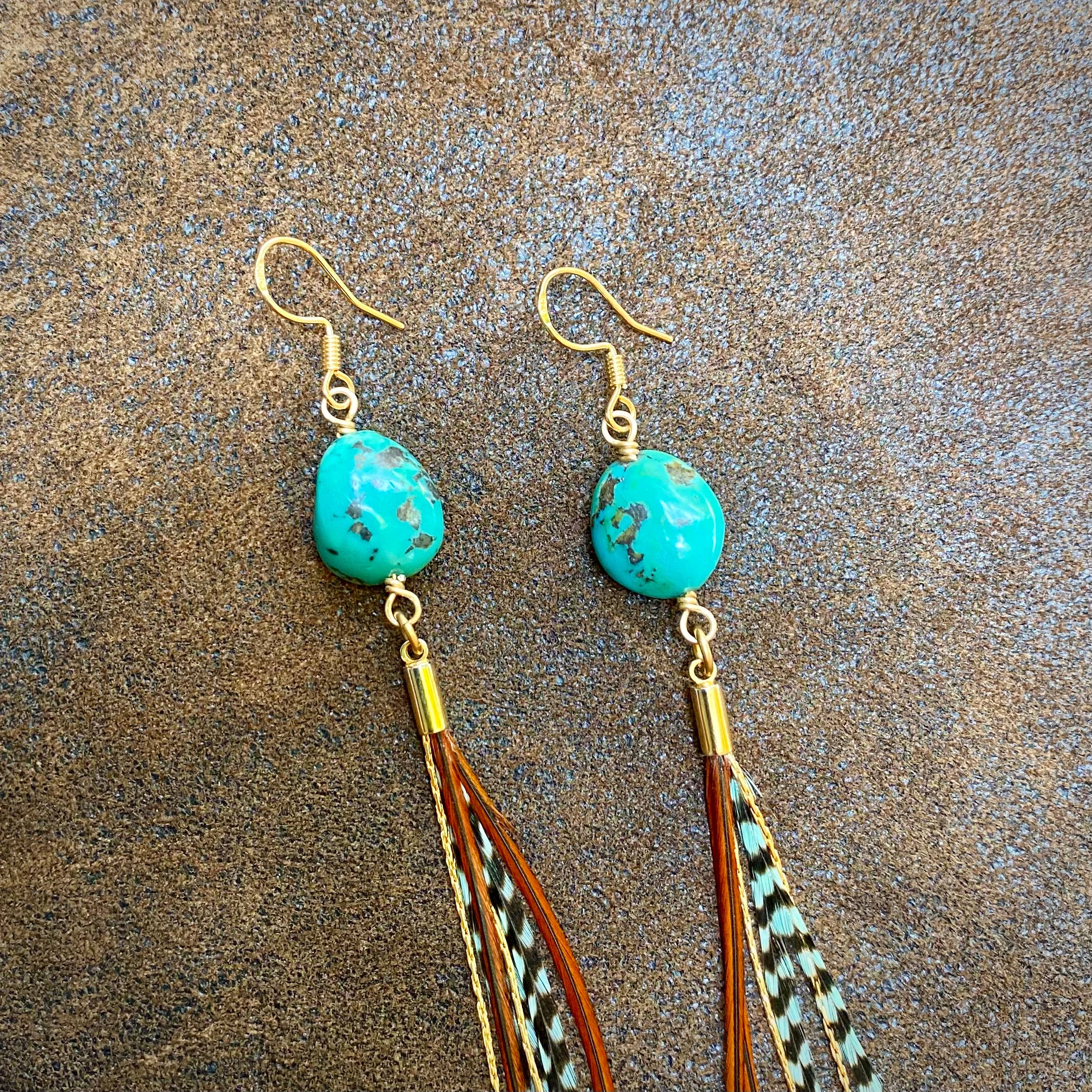 ASTALI Turquoise & Mini Feather Earrings—Teal Grizzly and Fawn feathers with gold hooks—are displayed on a textured brown surface.