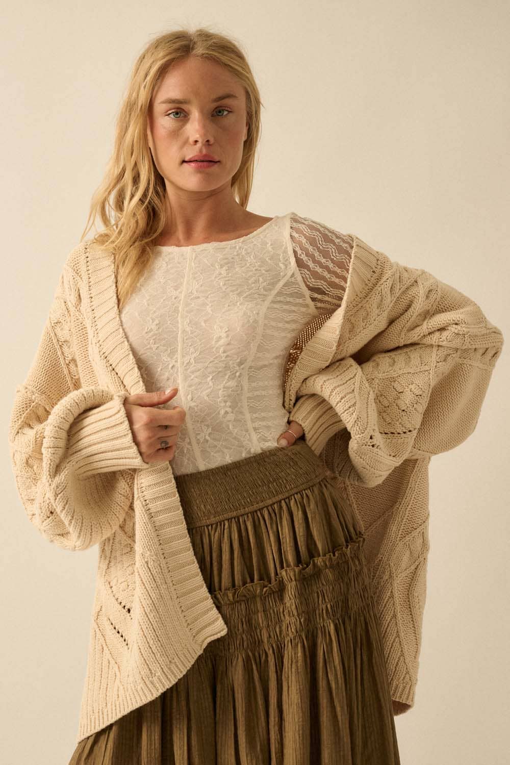 A woman with long blonde hair models the Promesa USA Cable-Knit Oversize Balloon-Sleeve Cardigan over a white lace top and brown pleated skirt, standing against a plain light background.