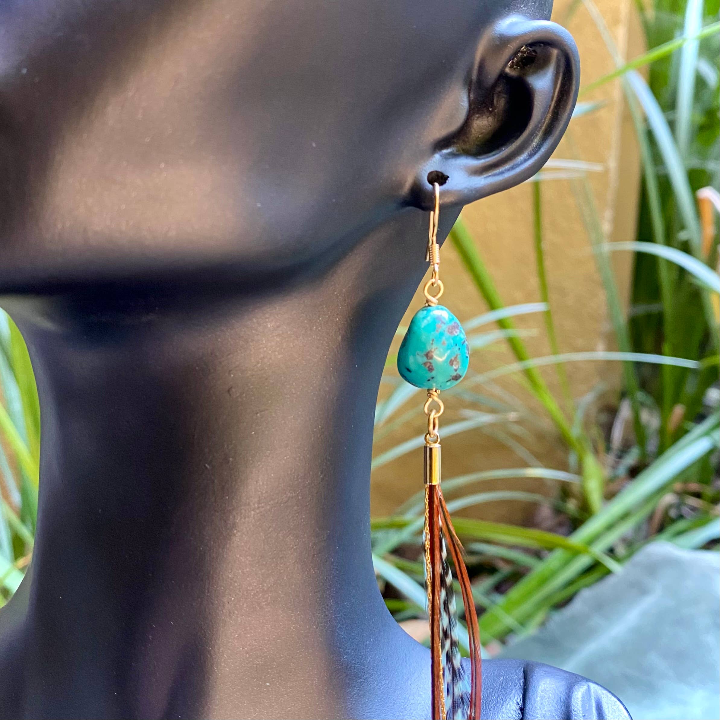 A close-up of ASTALI’s Turquoise & Mini Feather Earrings—Teal Grizzly & Fawn/Gold—on a mannequin’s ear, featuring handcrafted gold hardware and feather accents. Green plants and a tan wall are softly blurred in the background.
