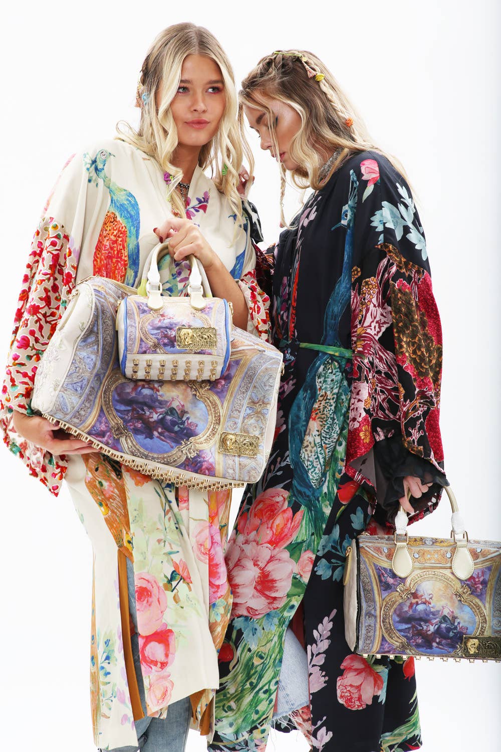 Two women wear Aratta’s Sweet Fantasy Kimono with burnout velvet sleeves, each holding ornate decorative bags featuring classical art prints and gold accents. One faces forward while the other looks down against a white background.