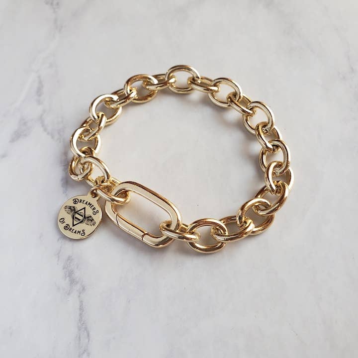 The Chunky Cable Chain Gold Plated Bracelet by Dreamers of Dreams features oval links and a round charm engraved with DREAMERS OF DREAMS and a star, shown on a white marble surface.
